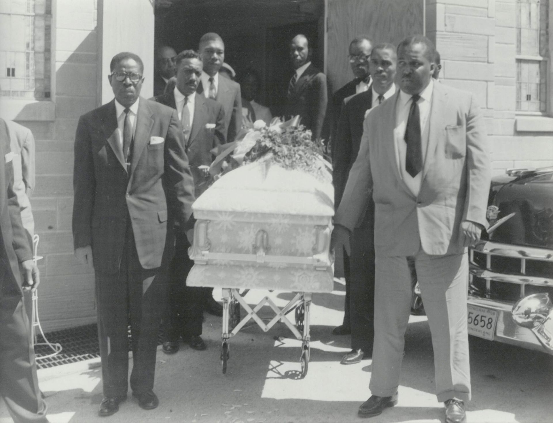 A framed black-and-white photograph shows a group of people in suits escorting a casket on a rolling stand outside a building.