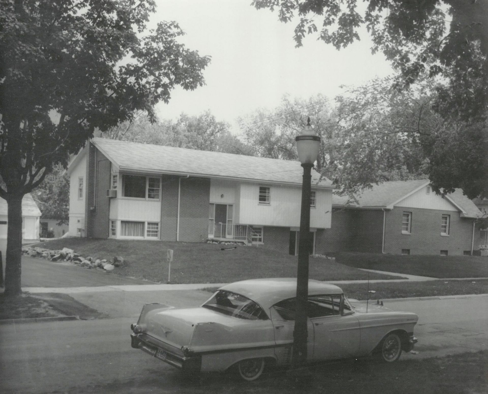 A black and white photograph of a vintage car parked on a suburban street in front of a house with trees.