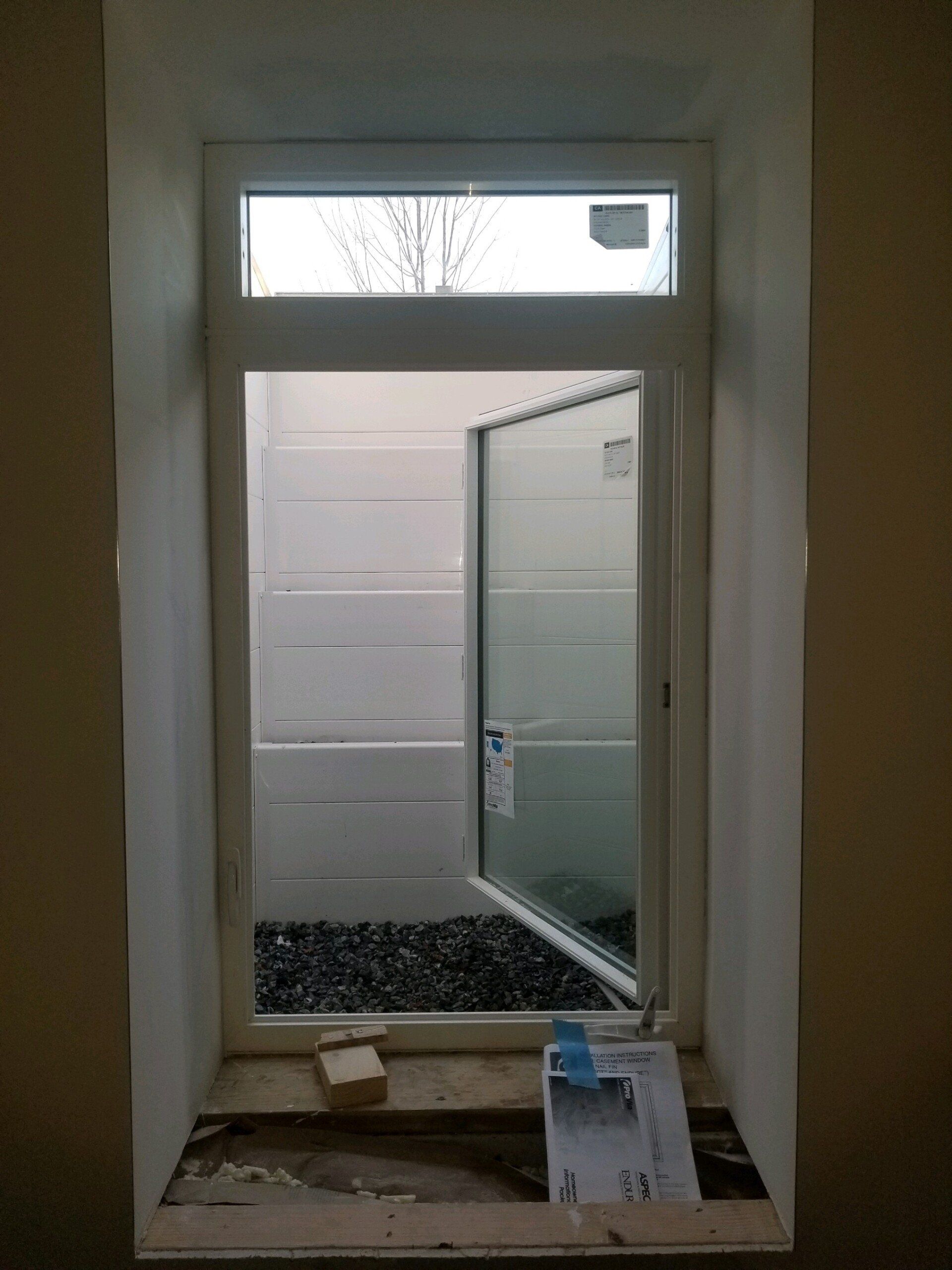A white window frame looking out at a basement window well filled with dark gravel, with construction debris on the sill.