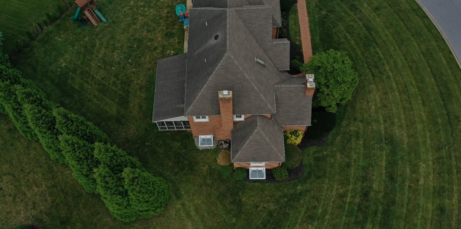 An aerial view of a brown brick home with a multi-gabled roof, surrounded by a large green lawn and tall privacy hedges.