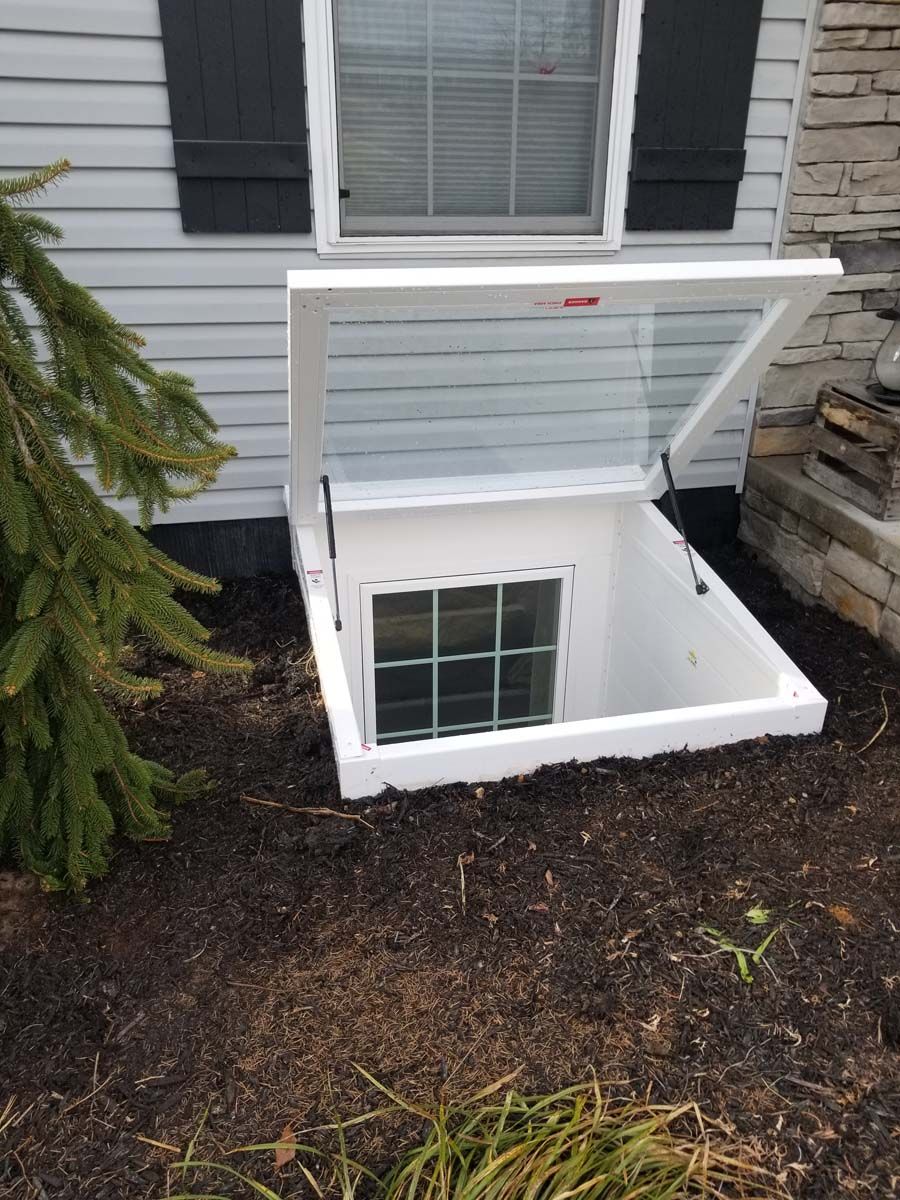 A white window well cover is propped open over a basement window embedded in the ground against a house exterior.