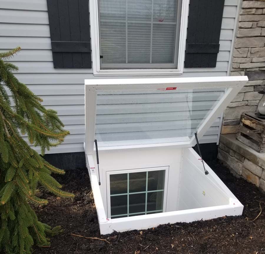 A white, hinged egress window well cover is propped open against the side of a gray-sided house with a basement window.