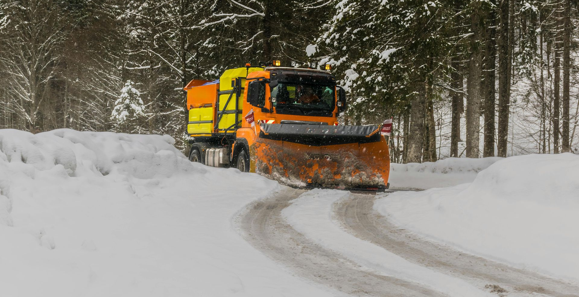 Snowplow clearing a snowy road with trees in background.
