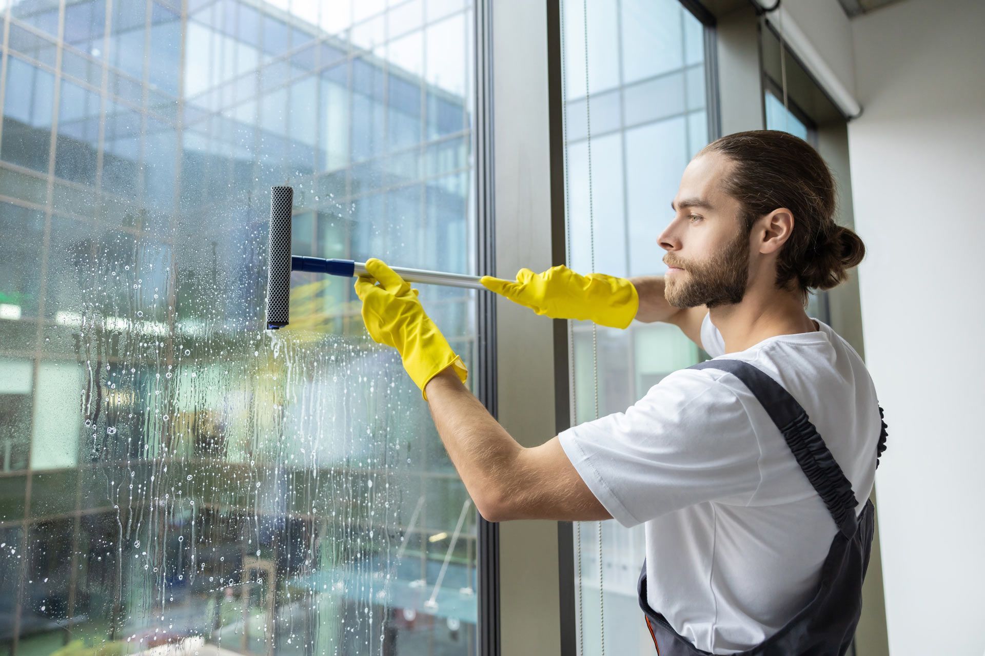 A person in a white shirt and gray overalls with yellow gloves uses a squeegee to clean a large window.