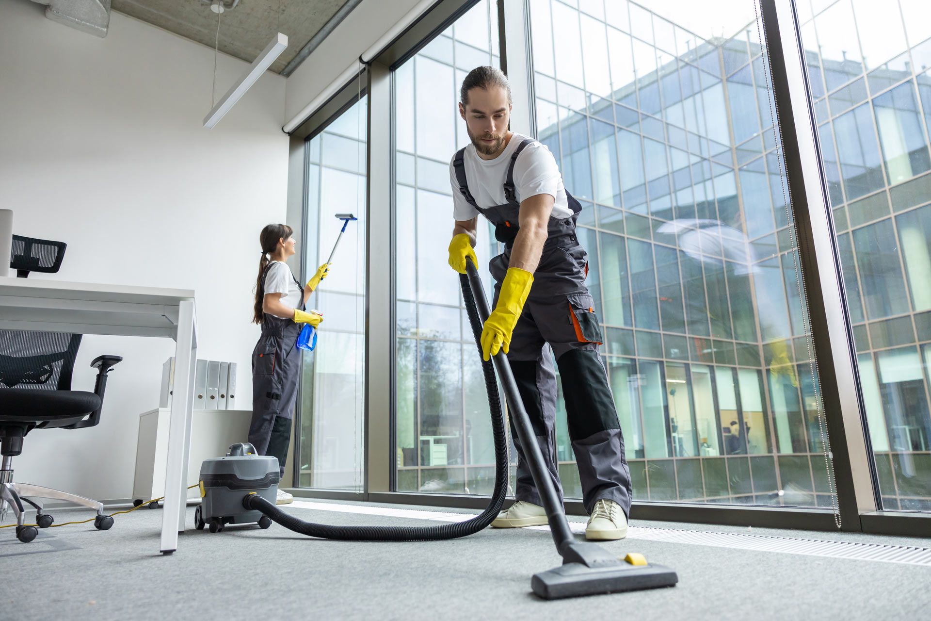 Two professional cleaners in grey jumpsuits clean a modern office with a vacuum cleaner and window squeegee.