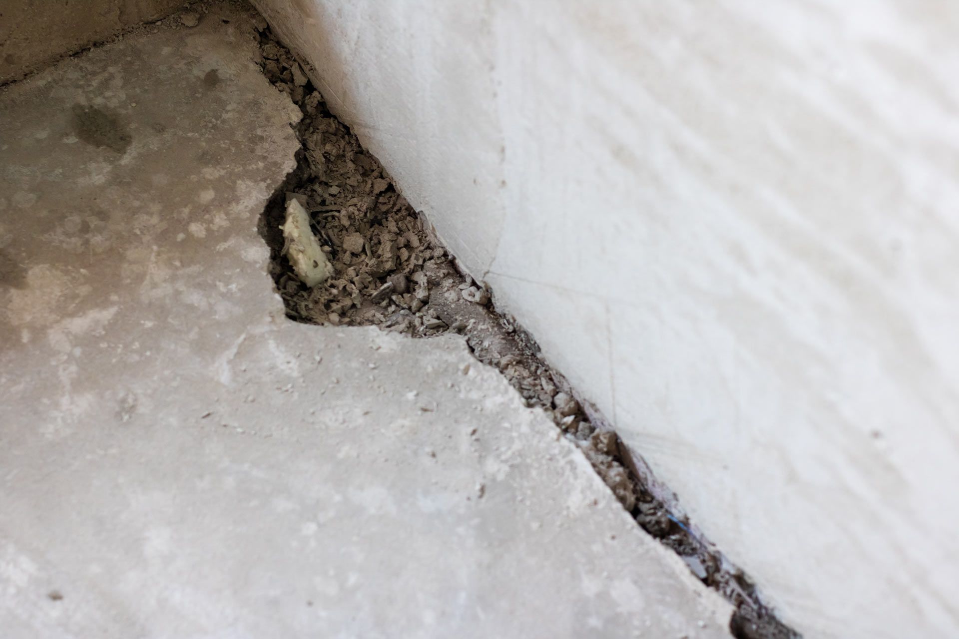 A cracked, light-colored concrete floor showing loose gravel debris along a wall-floor joint.