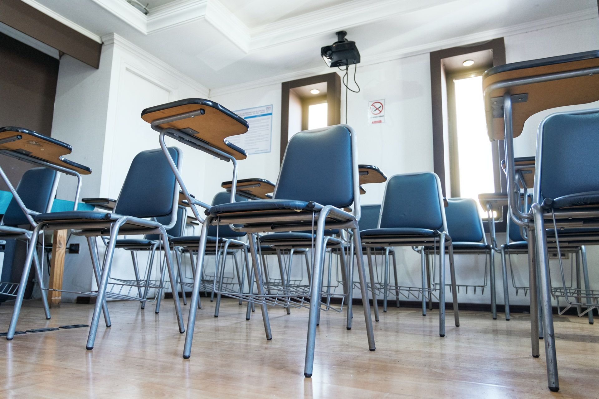 A classroom features multiple blue chairs with attached wooden desks arranged in rows facing a projector on the ceiling.