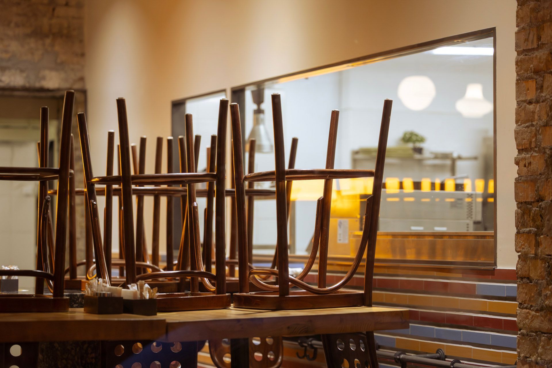 Wooden chairs sit upside down on tables in a rustic, brick-walled cafe setting, viewable through a large window.