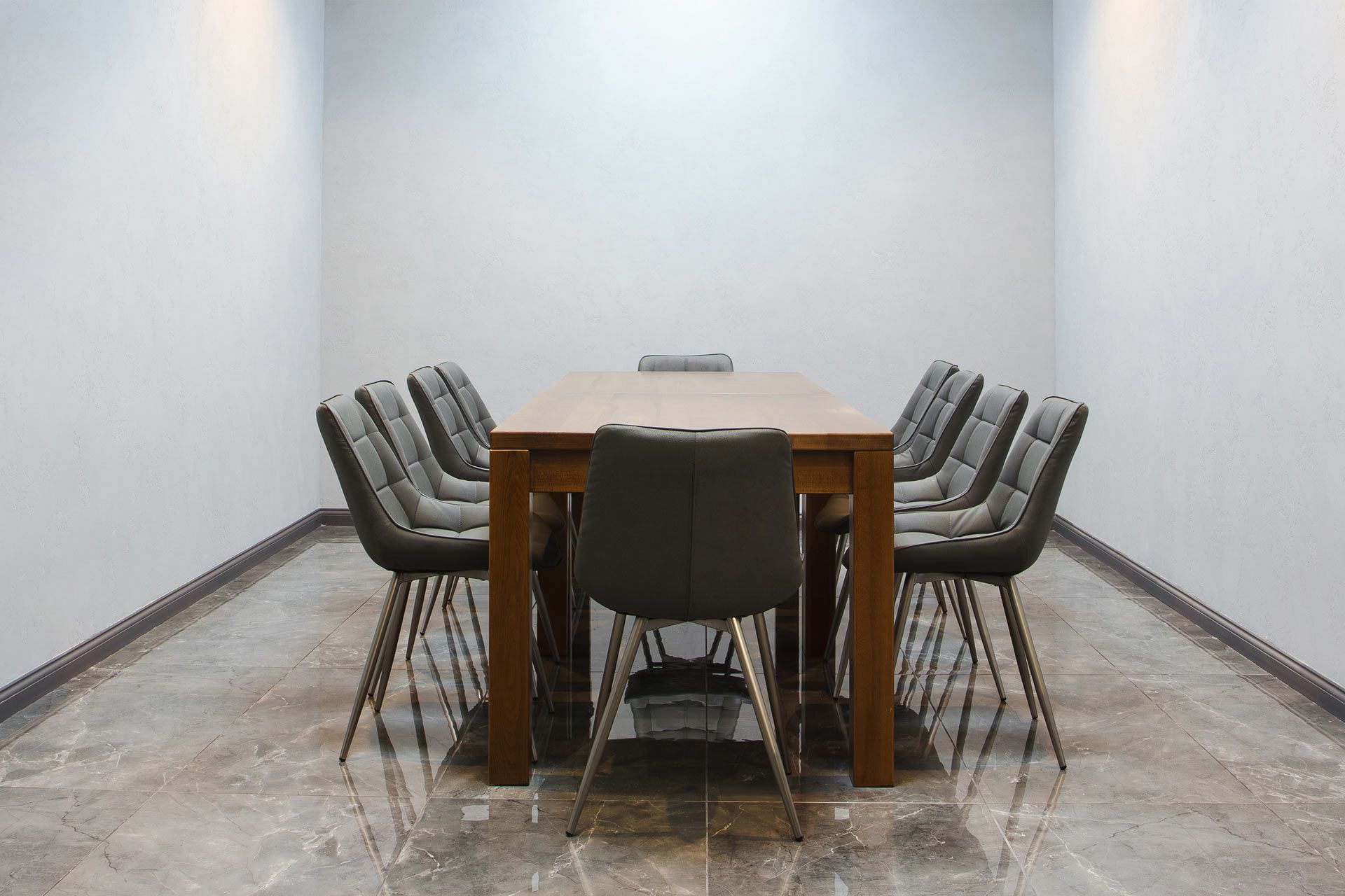 A modern, empty conference room with a large wooden table and ten gray upholstered chairs on a polished tile floor.