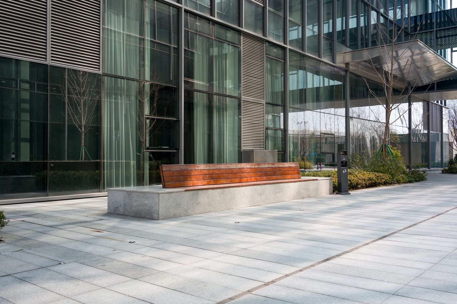 A long, wooden-topped bench with a grey stone base sits on a paved plaza in front of a modern glass office building.