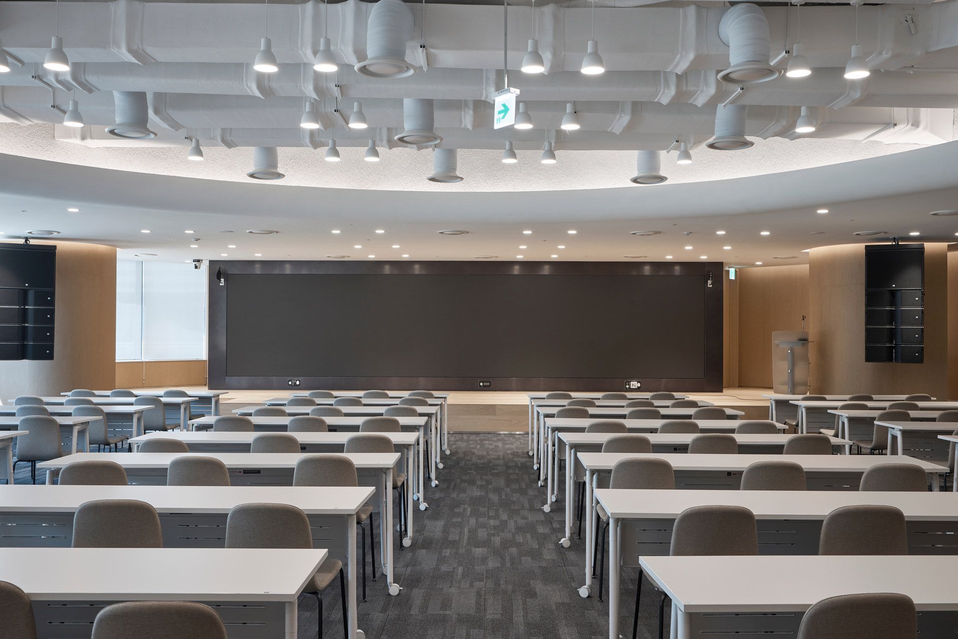 A wide, modern lecture hall with rows of white desks and chairs facing a large, dark digital screen on a curved wall.