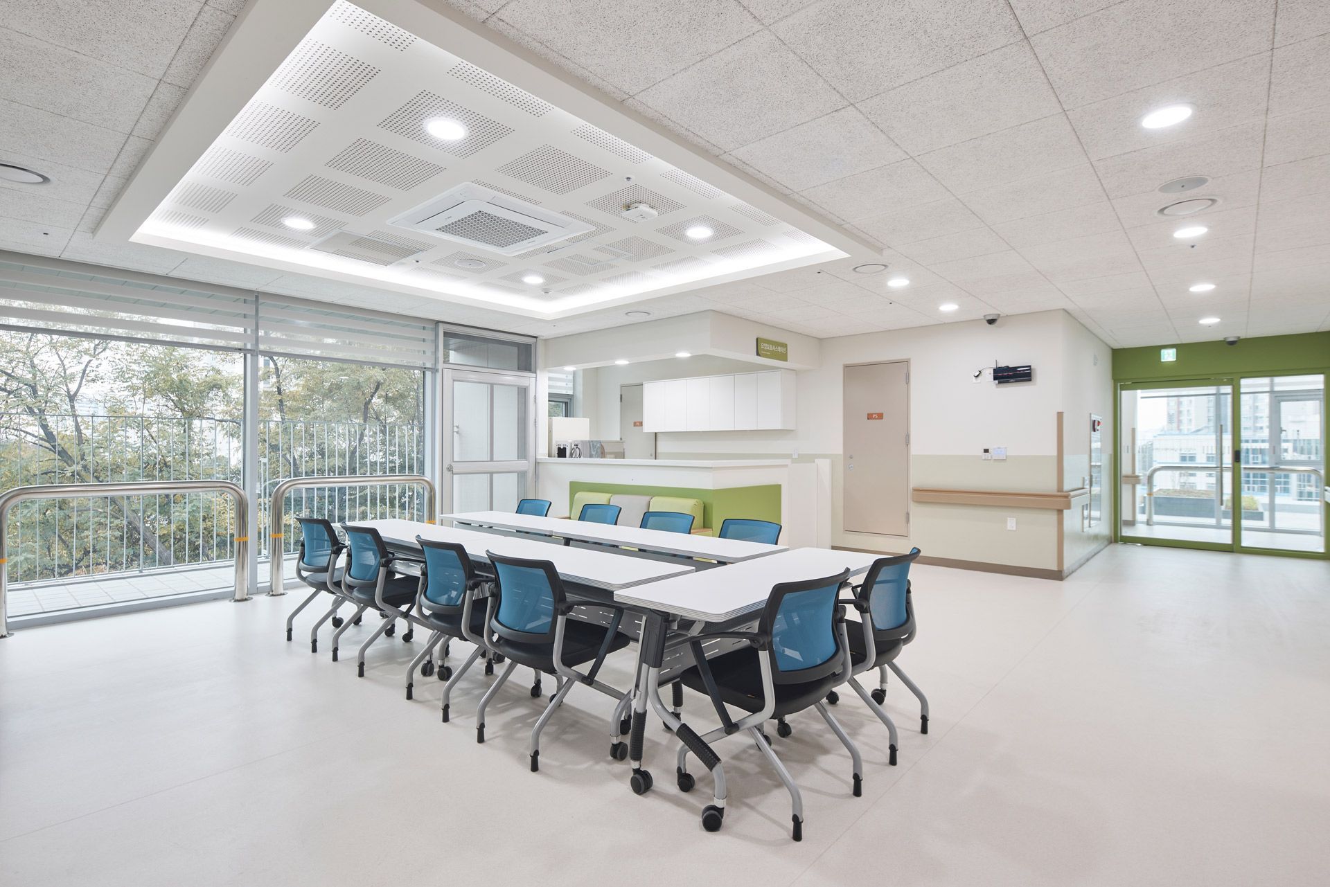A bright, modern meeting room with rows of blue chairs at white tables under a recessed, illuminated ceiling panel.