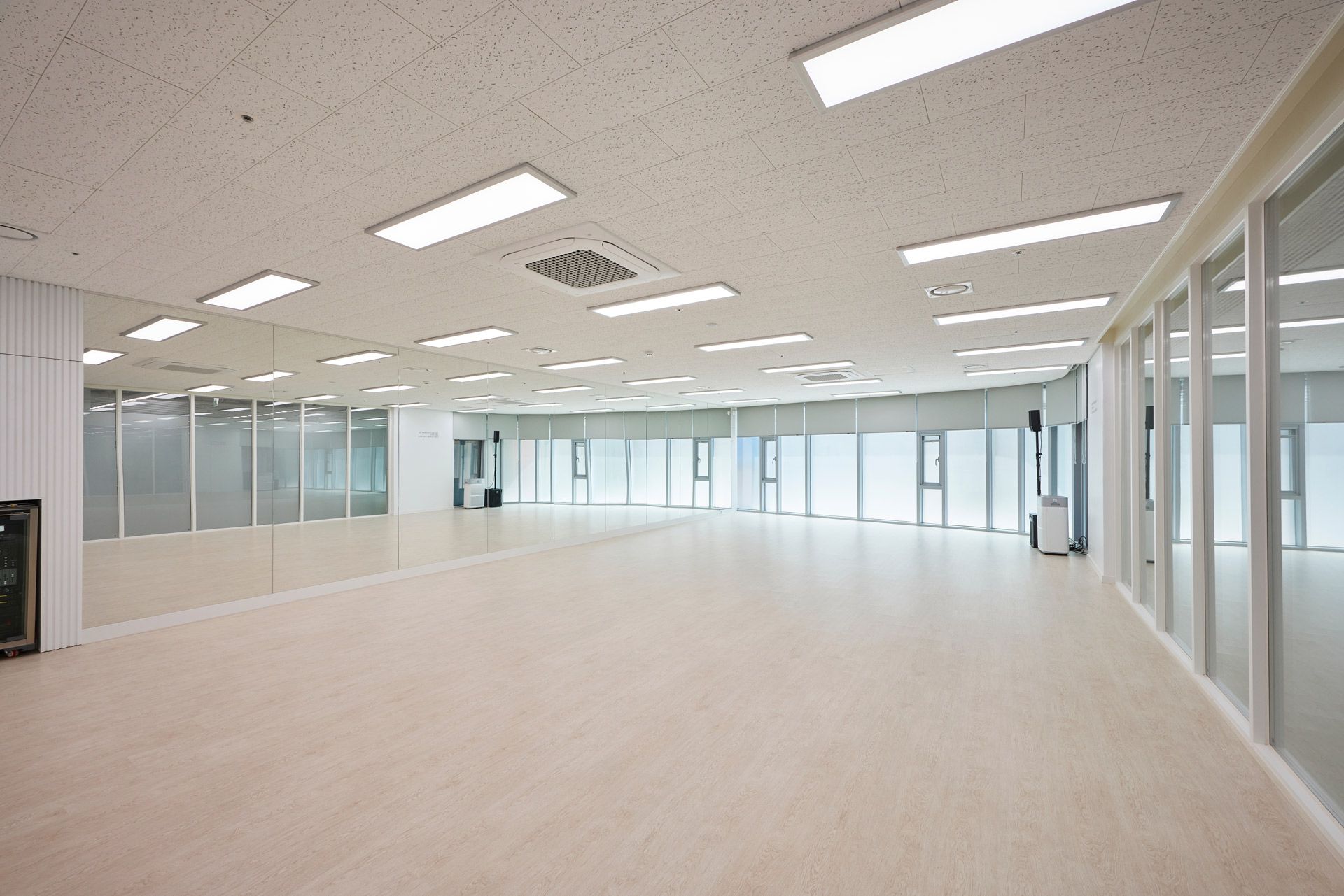 A spacious, empty dance studio with light wood flooring, mirrored walls, and large windows with blinds.