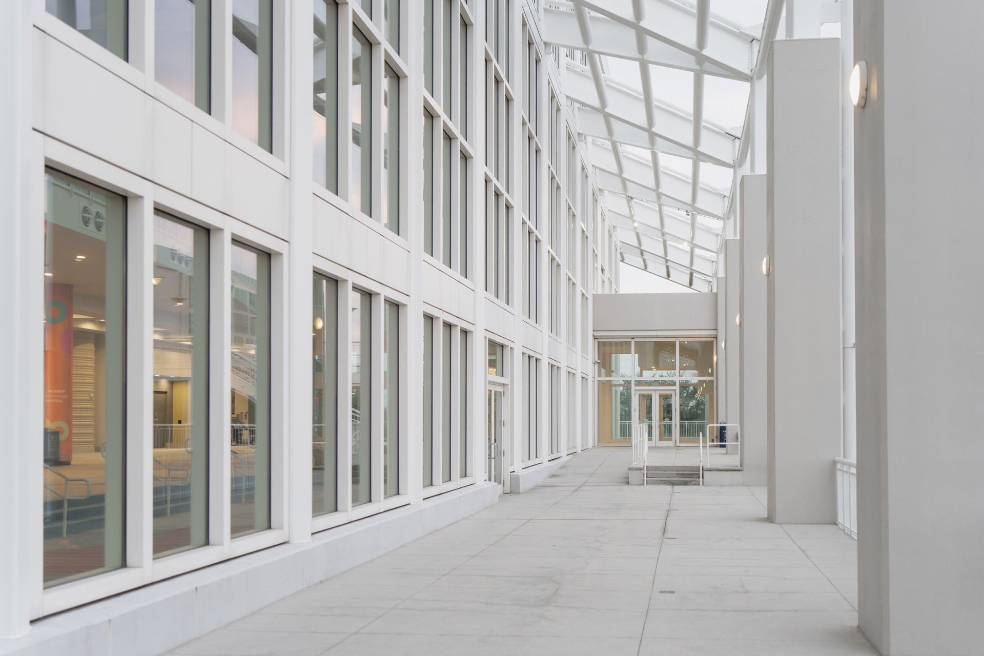 A modern, bright indoor hallway with white walls, floor-to-ceiling windows, and a sloped glass skylight ceiling.
