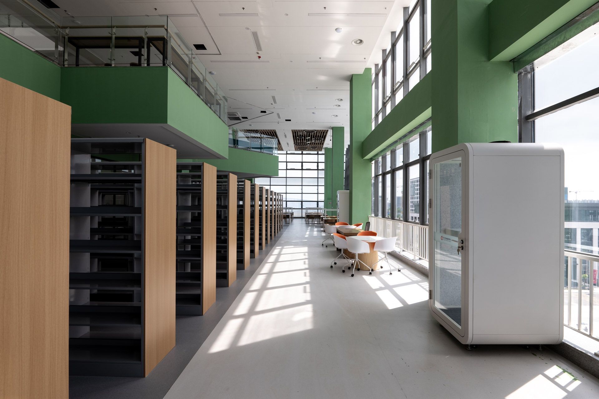 Modern library interior with green walls, rows of wooden bookshelves, study tables, and a white soundproof booth.