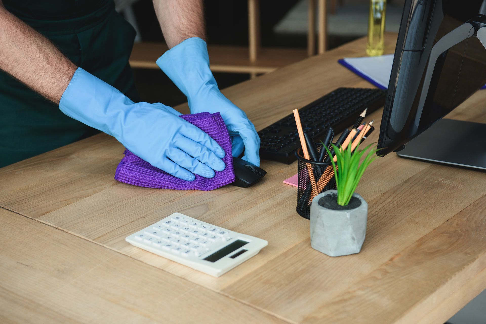 A person wearing blue gloves wipes a computer mouse on a wooden desk with a purple cloth, near a calculator and pen holder.