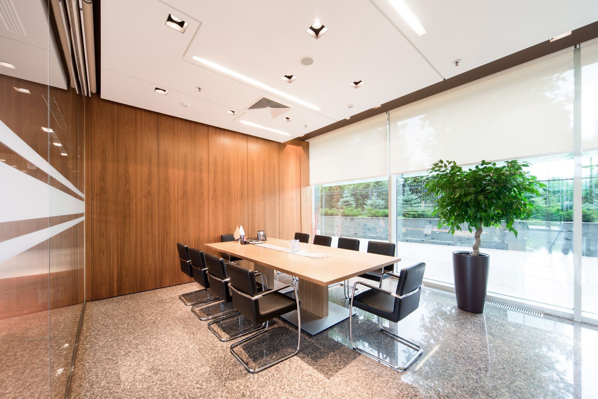 A modern conference room with a long wooden table, black office chairs, wood-paneled walls, and a potted tree.