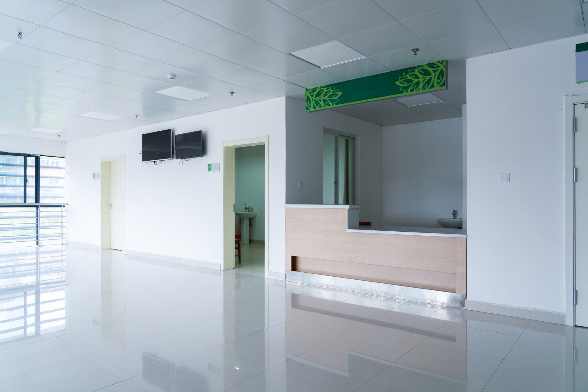A brightly lit, modern hospital reception area with white walls, a wooden desk, and a green decorative sign above.