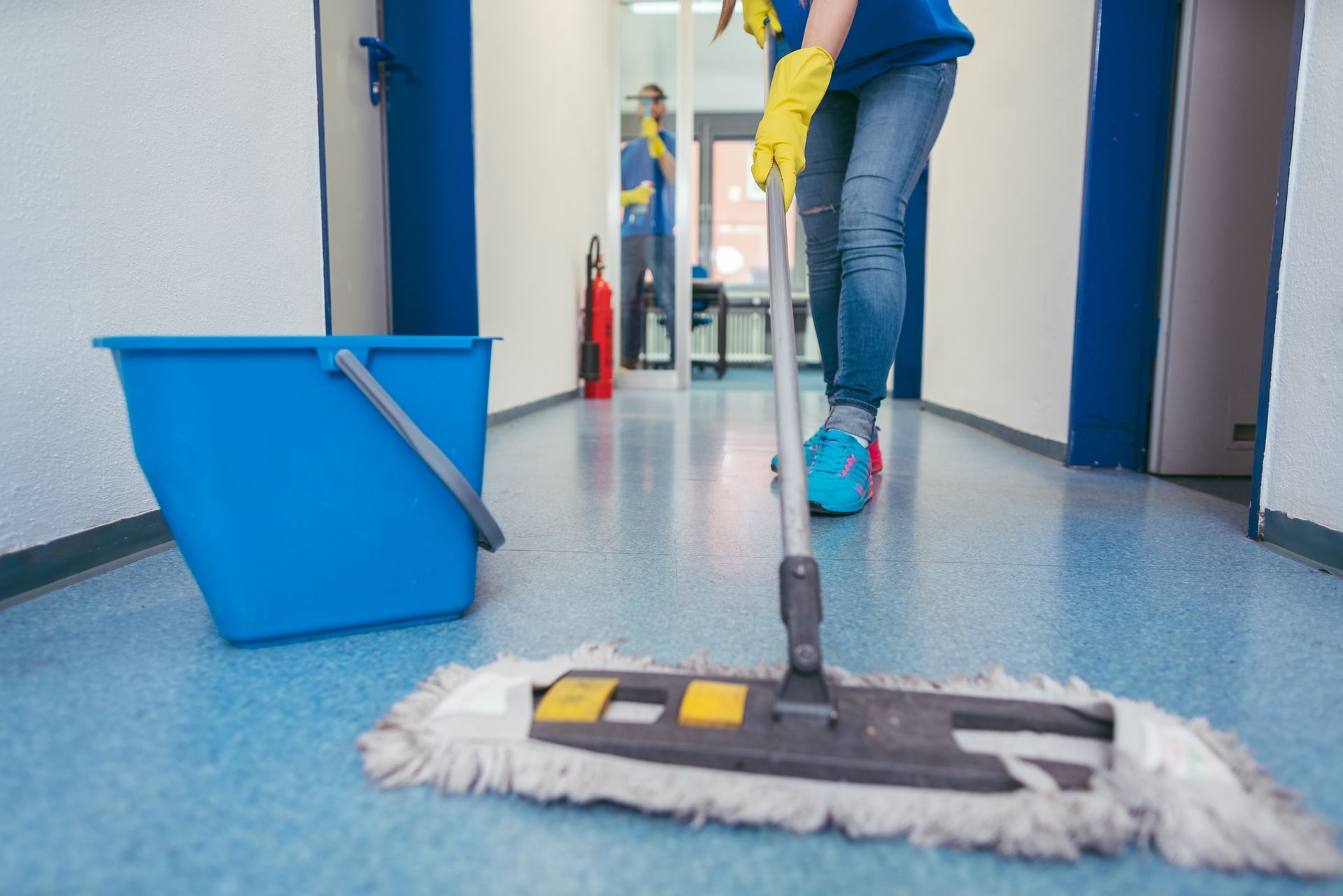 Person mopping a hallway floor with a flat mop, blue bucket nearby.
