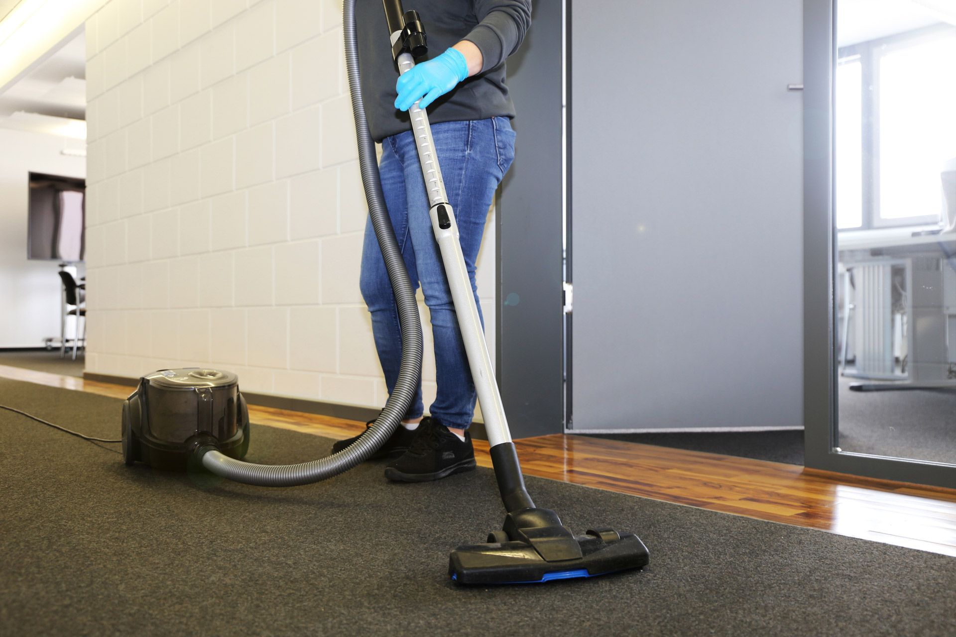 A person in blue gloves vacuums a dark office carpet in a hallway with white walls and a glass door.