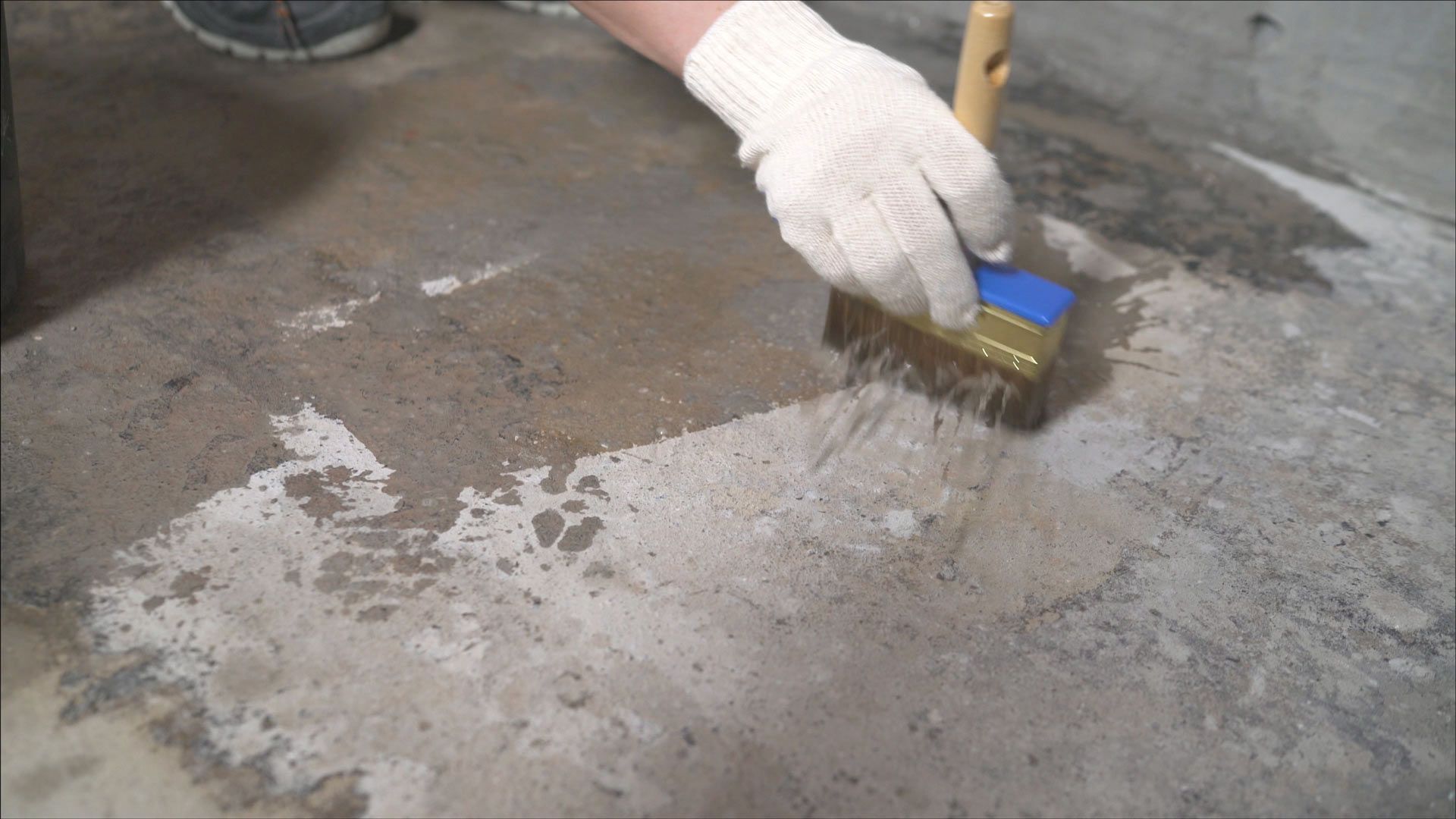 A gloved hand uses a brush to apply a liquid primer or sealant onto a rough concrete floor.