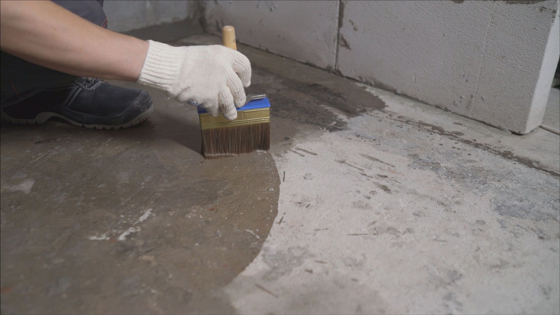 A person wearing a white glove uses a brush to apply a liquid primer to a concrete floor near a masonry wall.