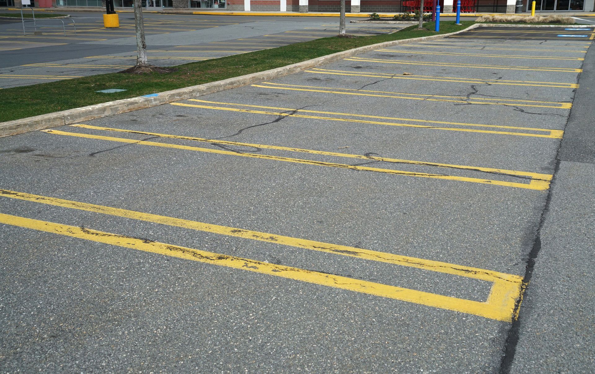 An asphalt parking lot with empty spaces outlined in worn, yellow paint, adjacent to a grassy median and a storefront.