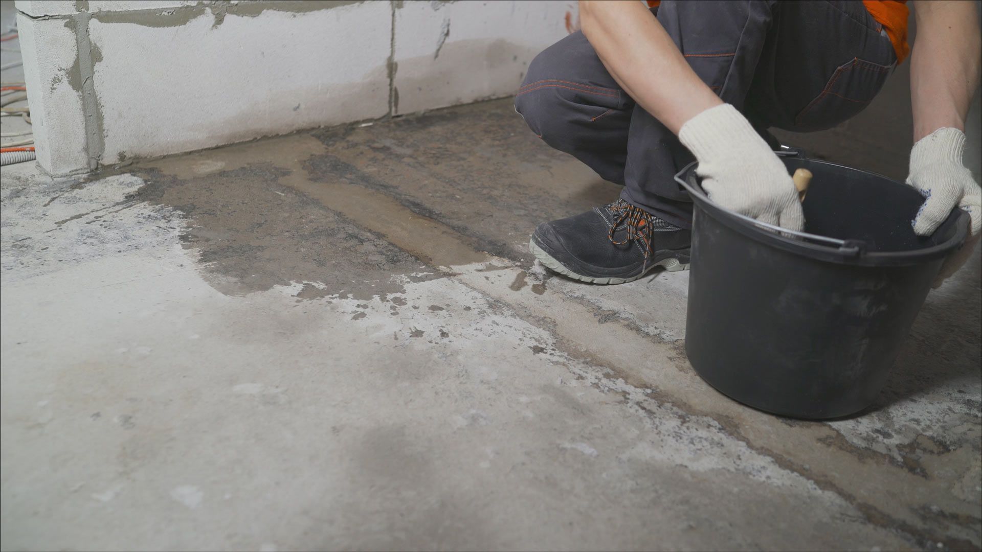 A person wearing work gloves kneels on a concrete floor, using a bucket to apply mortar to a seam between floor sections.
