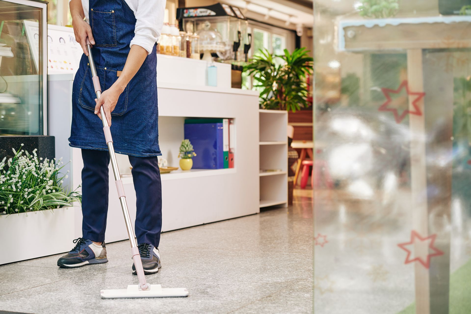 Person mopping a gray floor inside a cafe, wearing an apron.