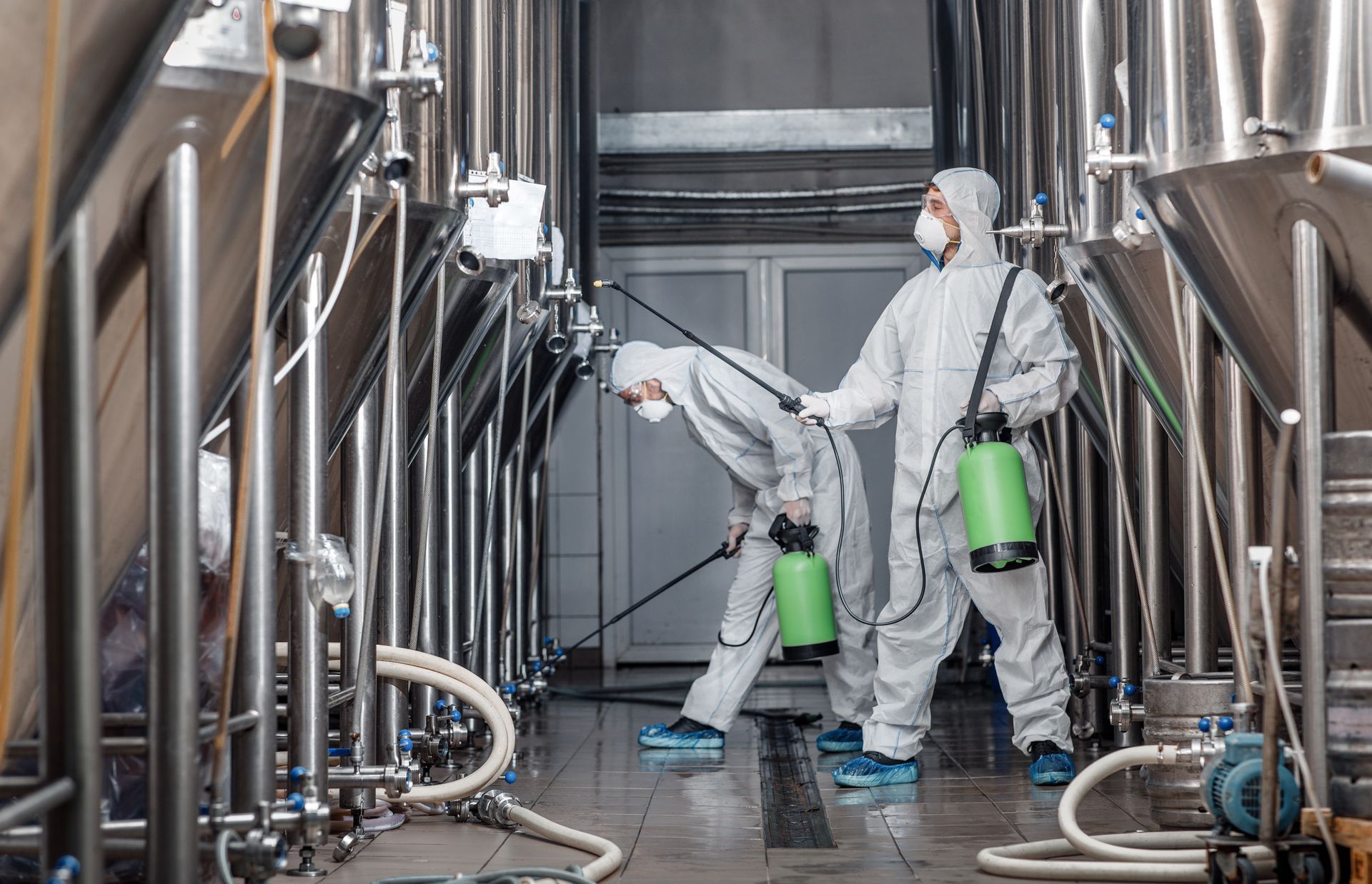 Two workers in protective suits spraying disinfectant in a brewery, among metal tanks and pipes.