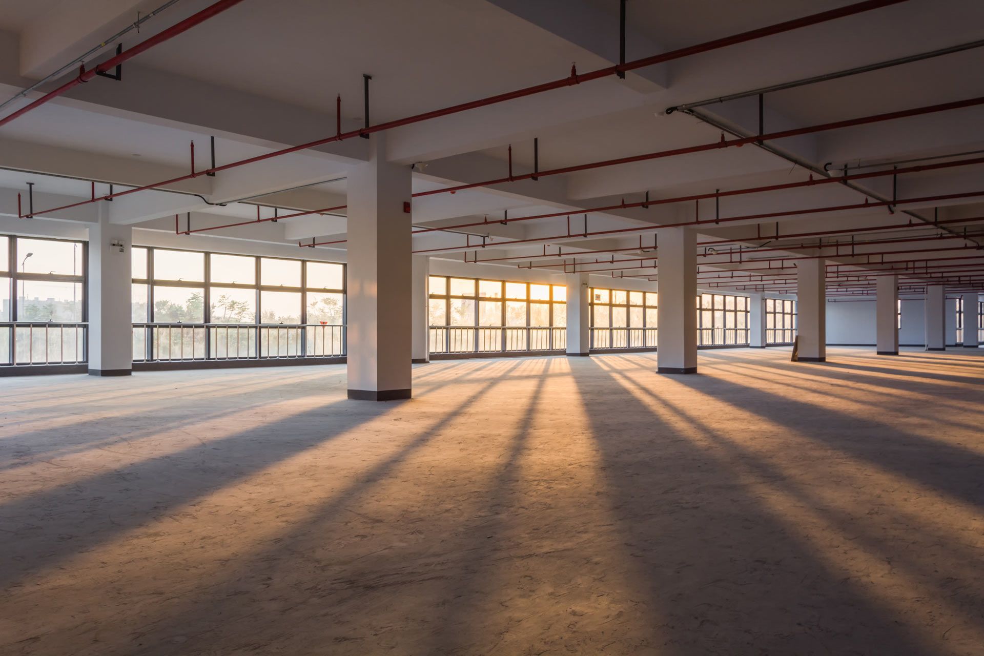 Sunlight streams through large windows into an empty, industrial-style concrete room with exposed pipes on the ceiling.