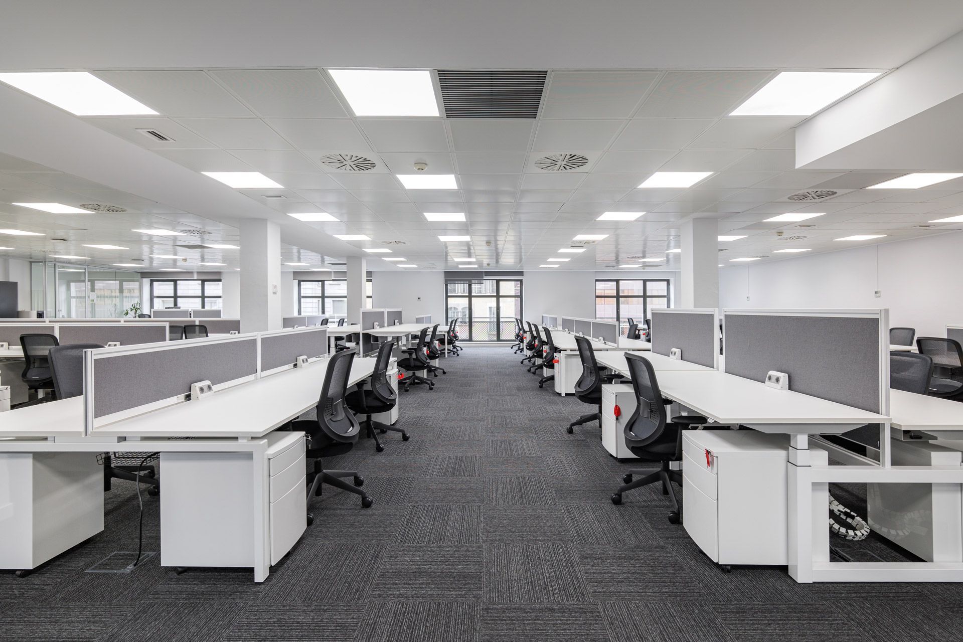 Modern, empty open-plan office with white desks, grey partitions, and rolling chairs on a dark carpeted floor.