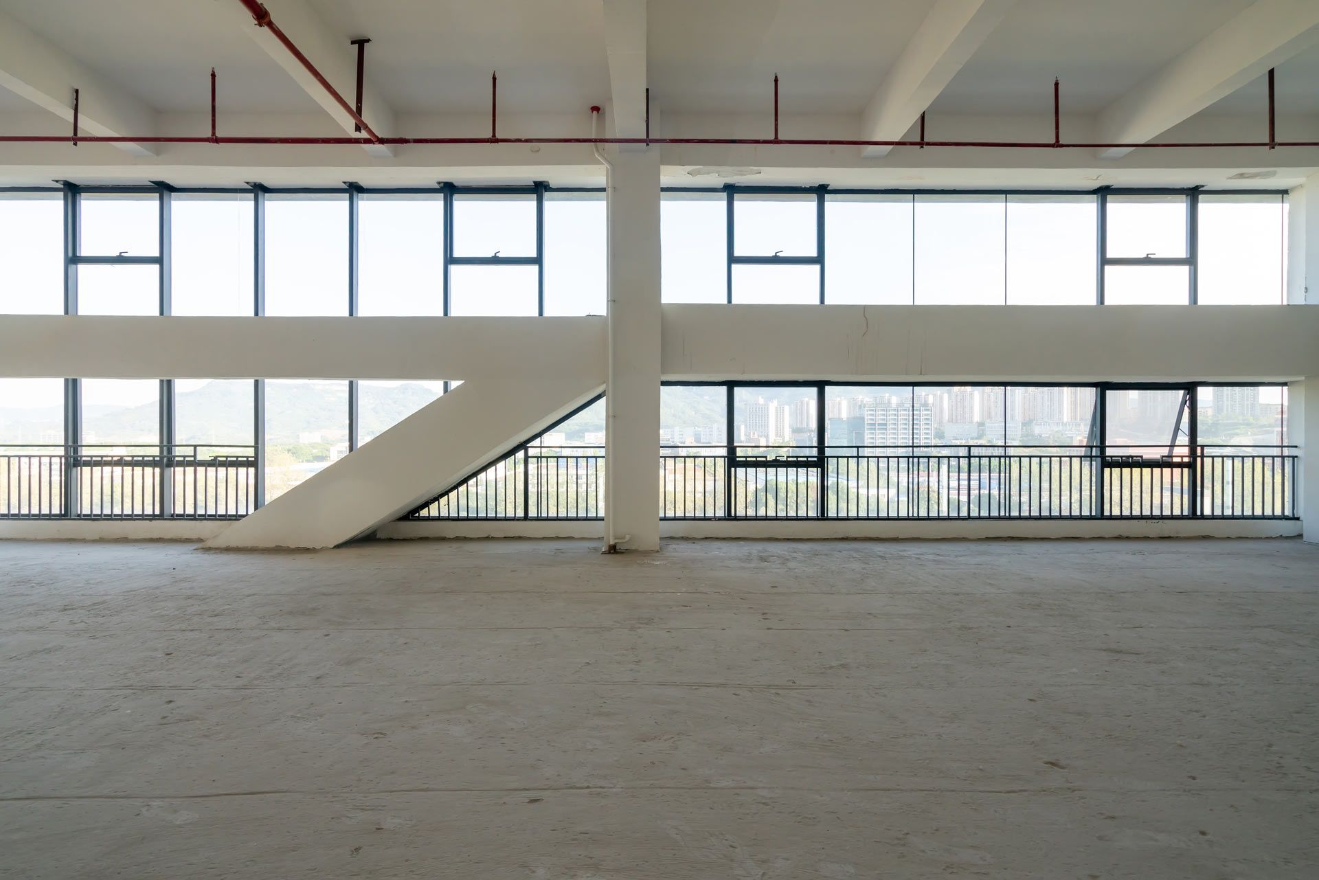 An empty, modern office space with concrete floors, large windows, and an angular architectural beam.