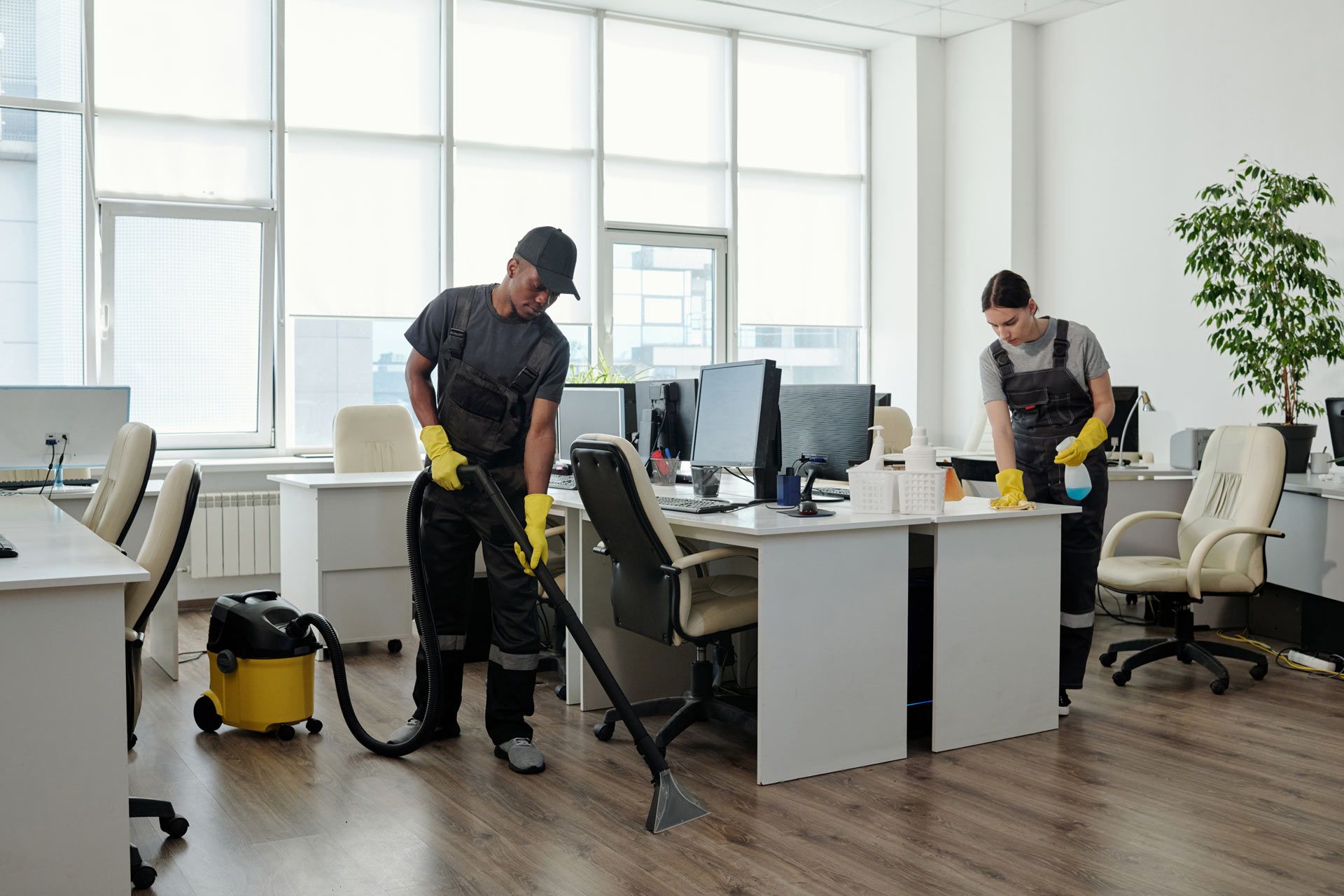 Two professional cleaners in dark uniforms and yellow gloves clean a bright, modern office with a vacuum and spray cleaner.