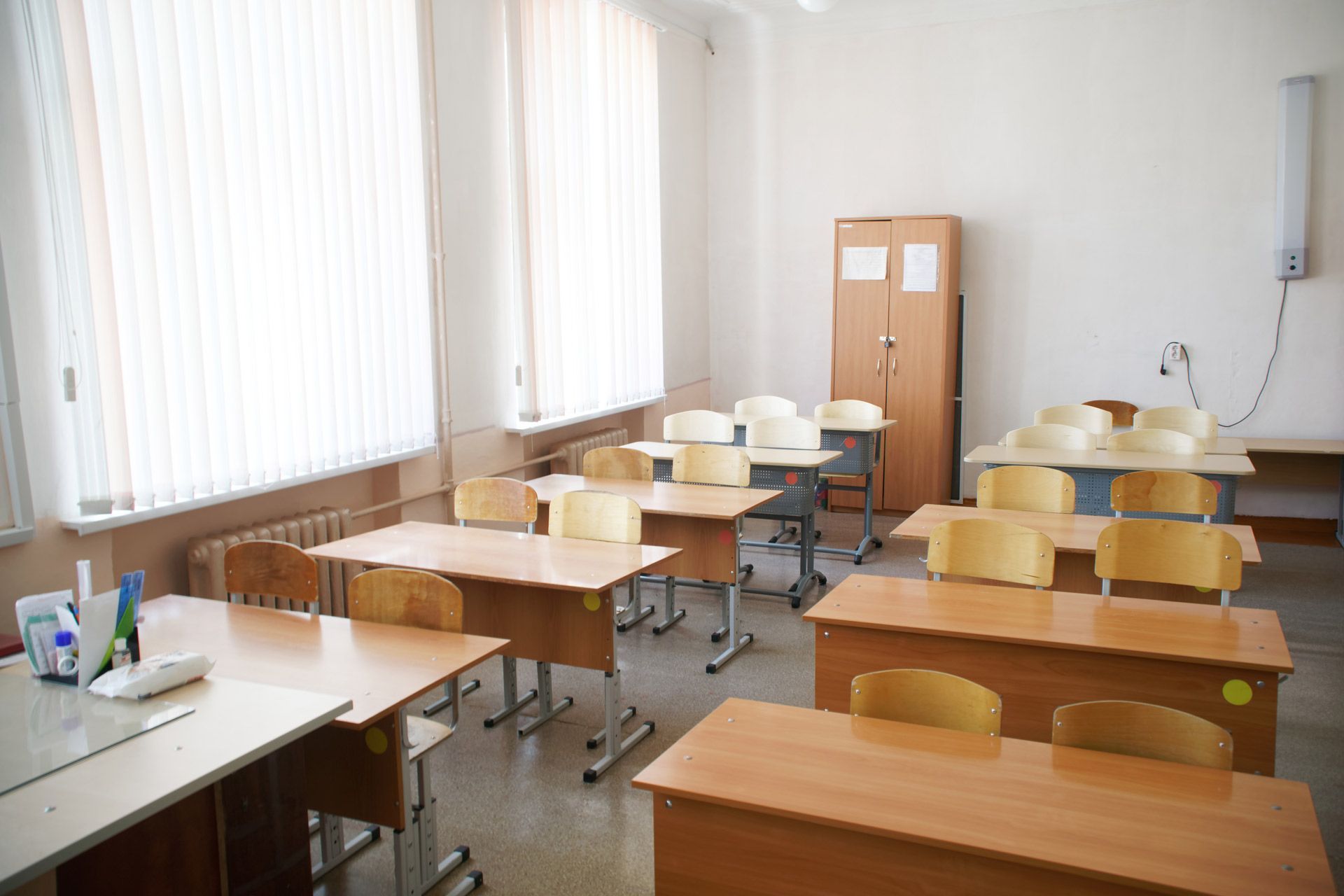 Empty classroom with rows of wooden desks and chairs, white walls, and window blinds.