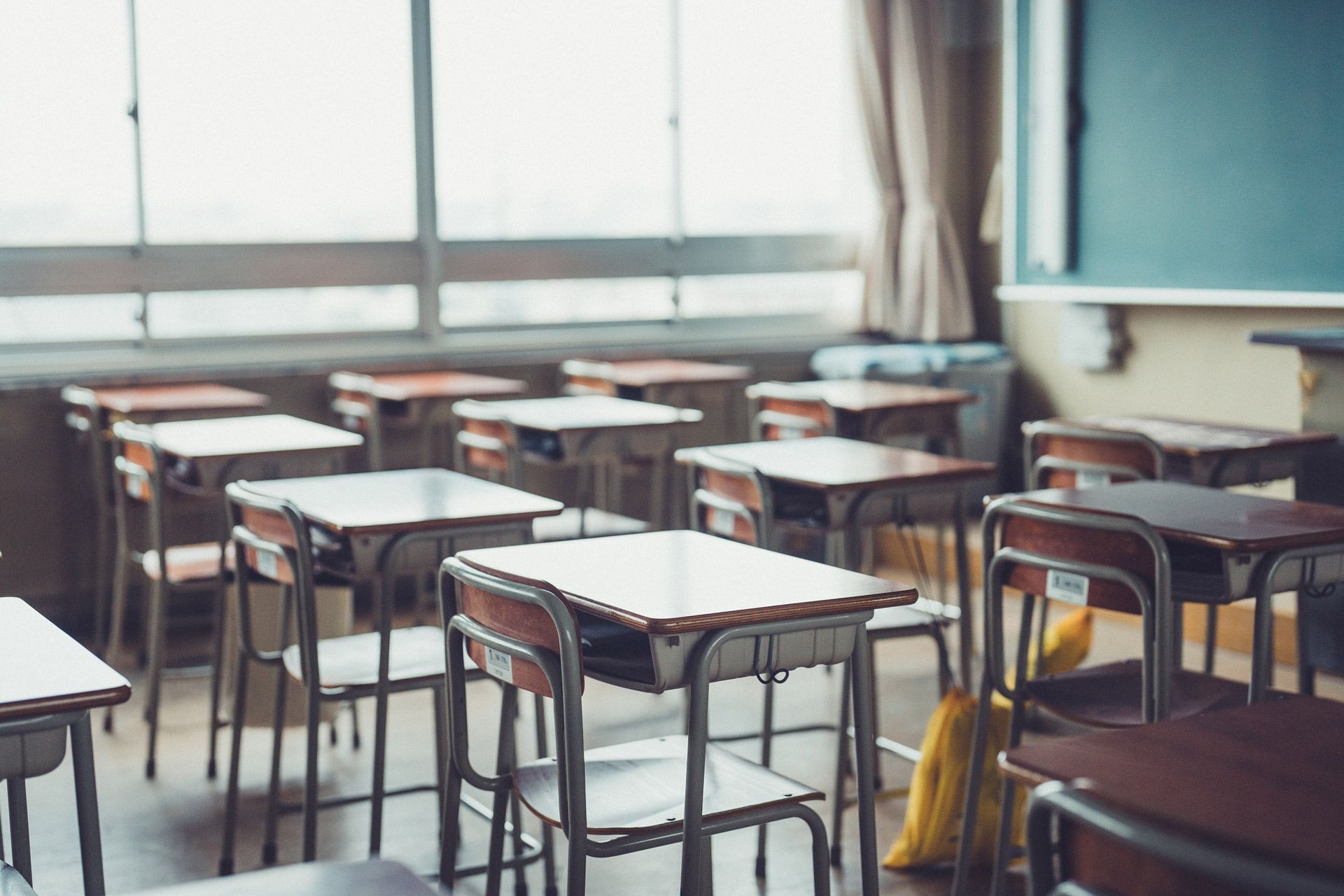 An empty classroom with rows of wooden student desks and chairs facing a chalkboard in front of large windows.