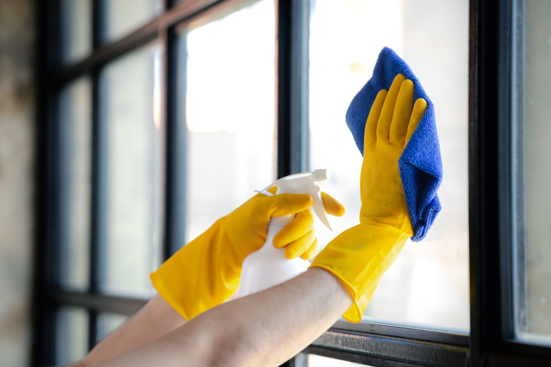 Hands in bright yellow rubber gloves use a blue cloth and spray bottle to clean a pane of a black-framed window.