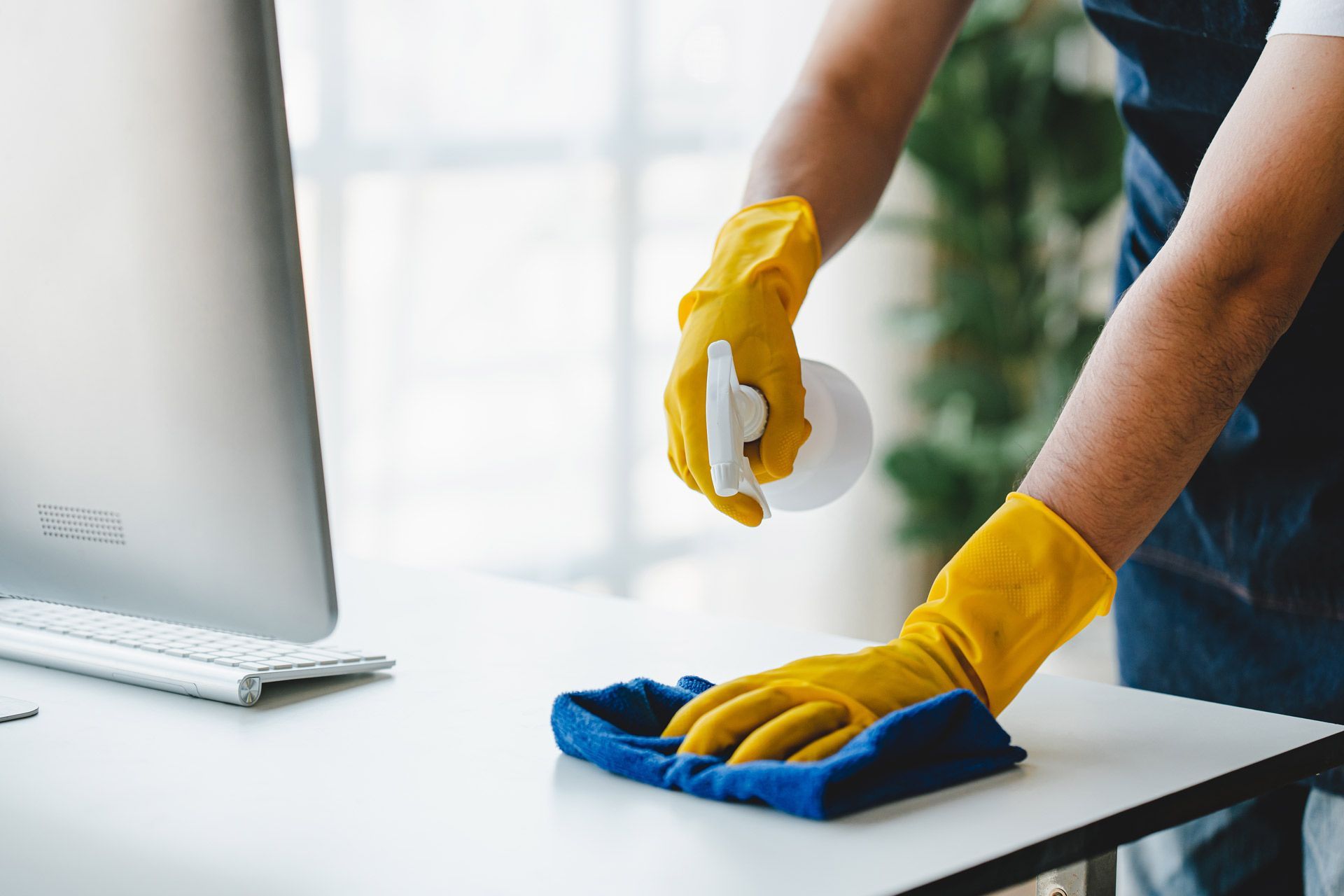 A person wearing yellow rubber gloves wipes a white desk with a blue cloth while holding a spray bottle.