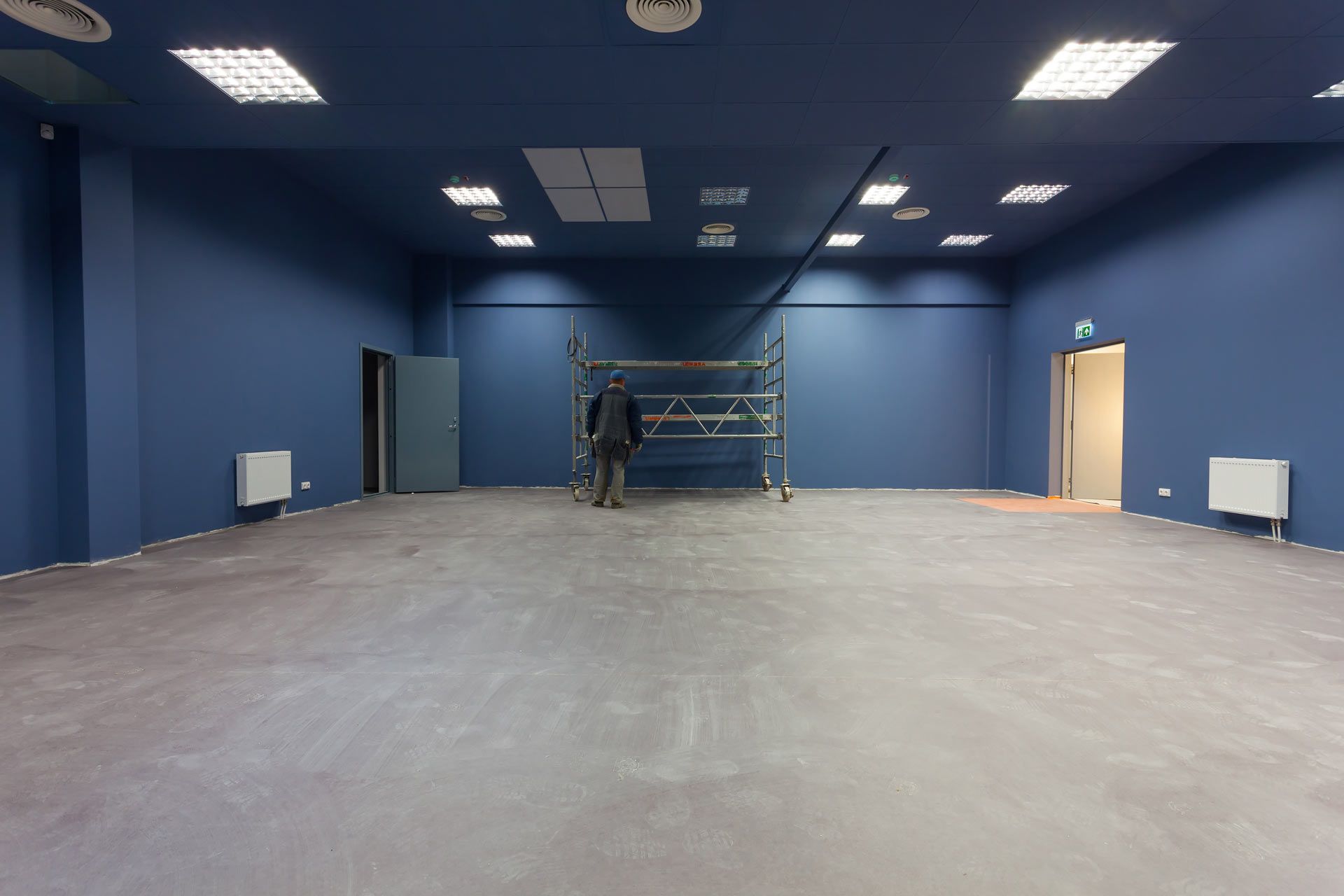 A person stands near scaffolding in a large, empty room with deep blue walls and gray flooring under bright ceiling lights.