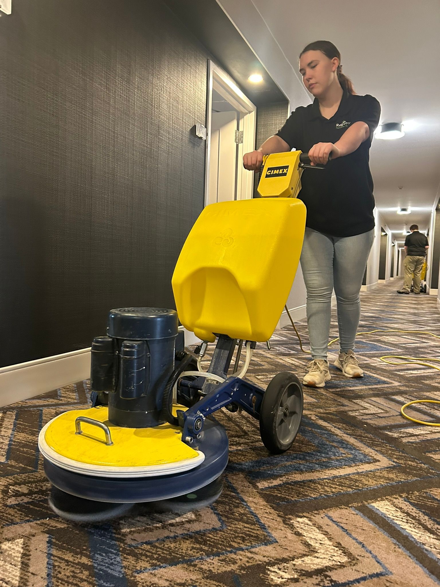 Woman using a yellow floor cleaning machine to clean a carpeted hallway.