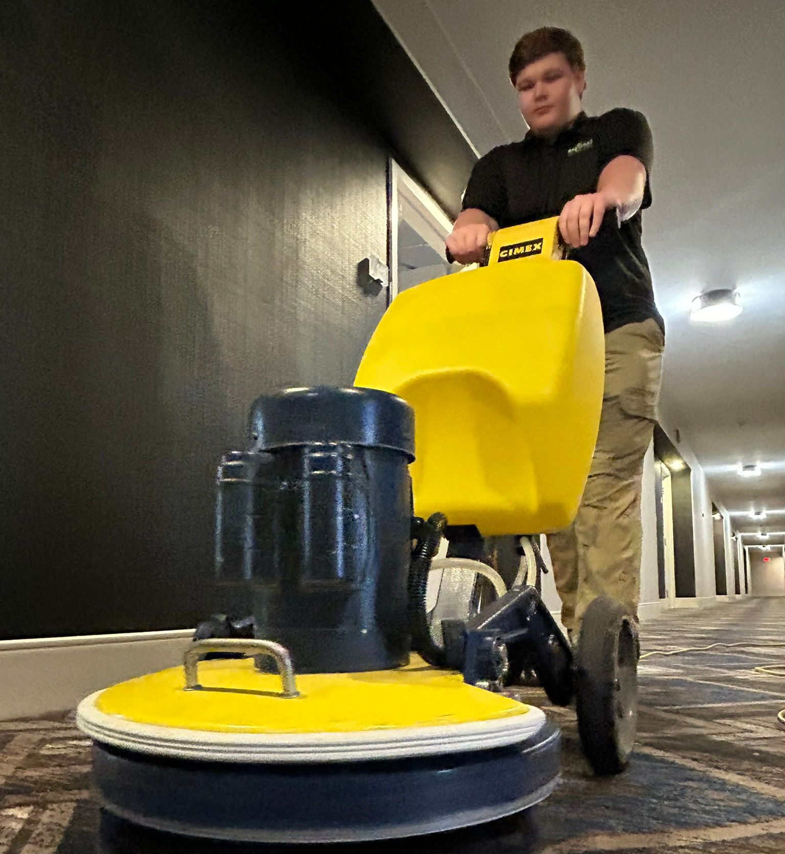 Person vacuuming a gray carpeted floor in a room with chairs and green walls. They wear orange and blue work clothes.