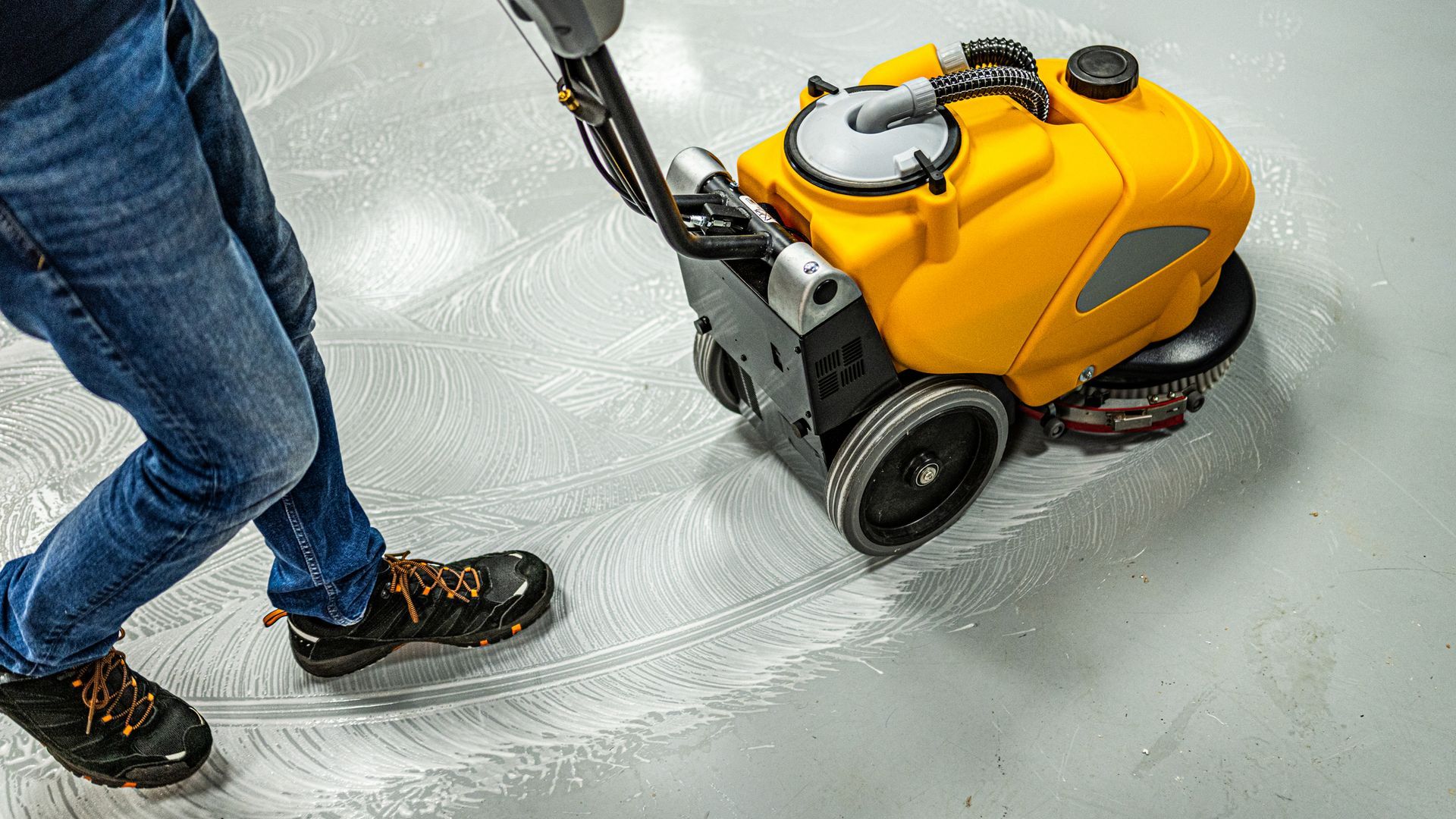 Person in blue jeans using a yellow floor scrubber machine on a white floor.