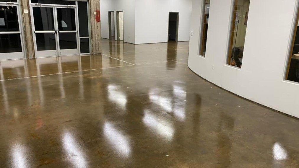 Person using floor buffer in a brightly lit office; another person cleans furniture.