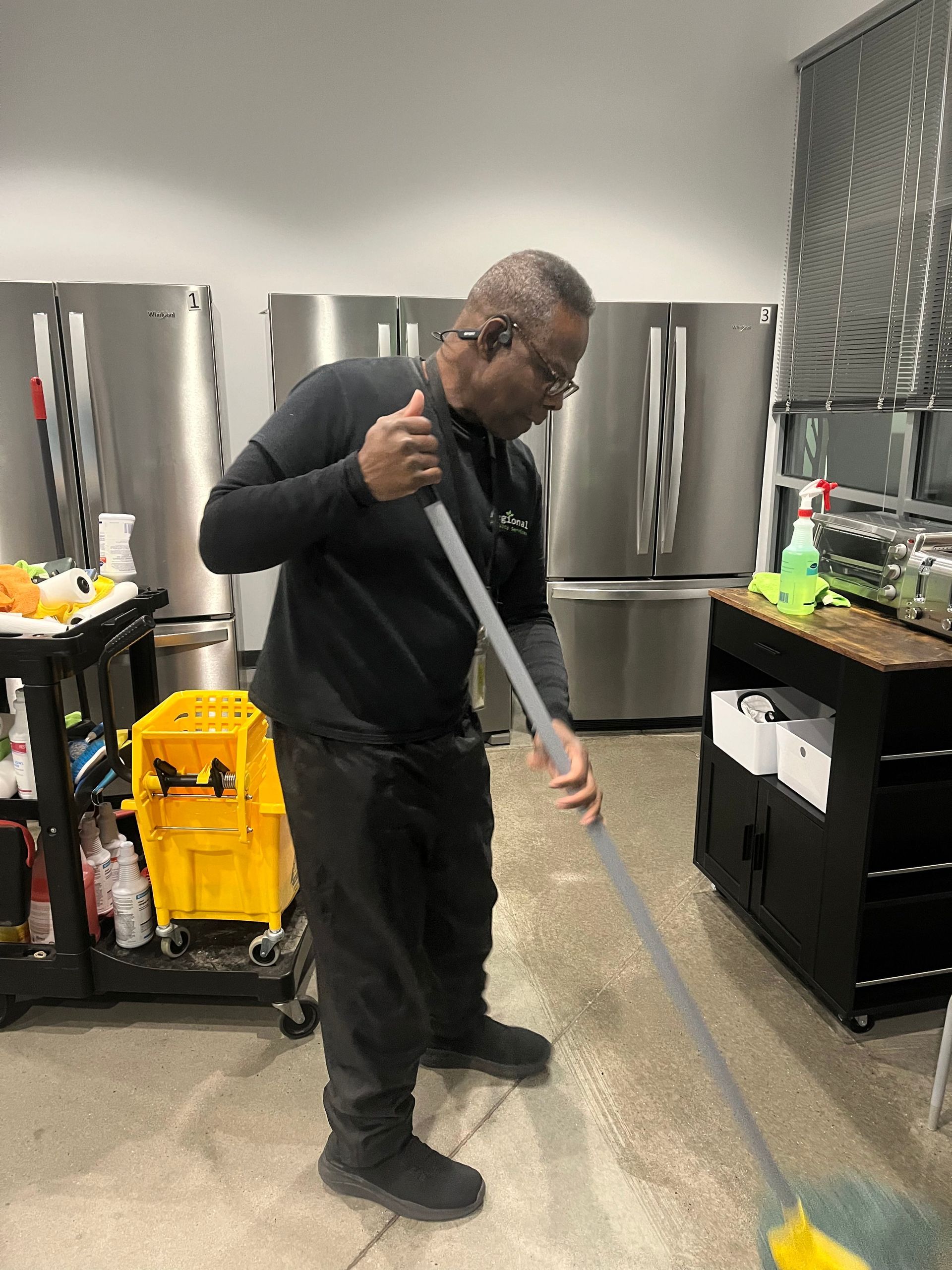 Person mopping a concrete floor in a commercial kitchen; stainless steel refrigerators and cleaning supplies are visible.