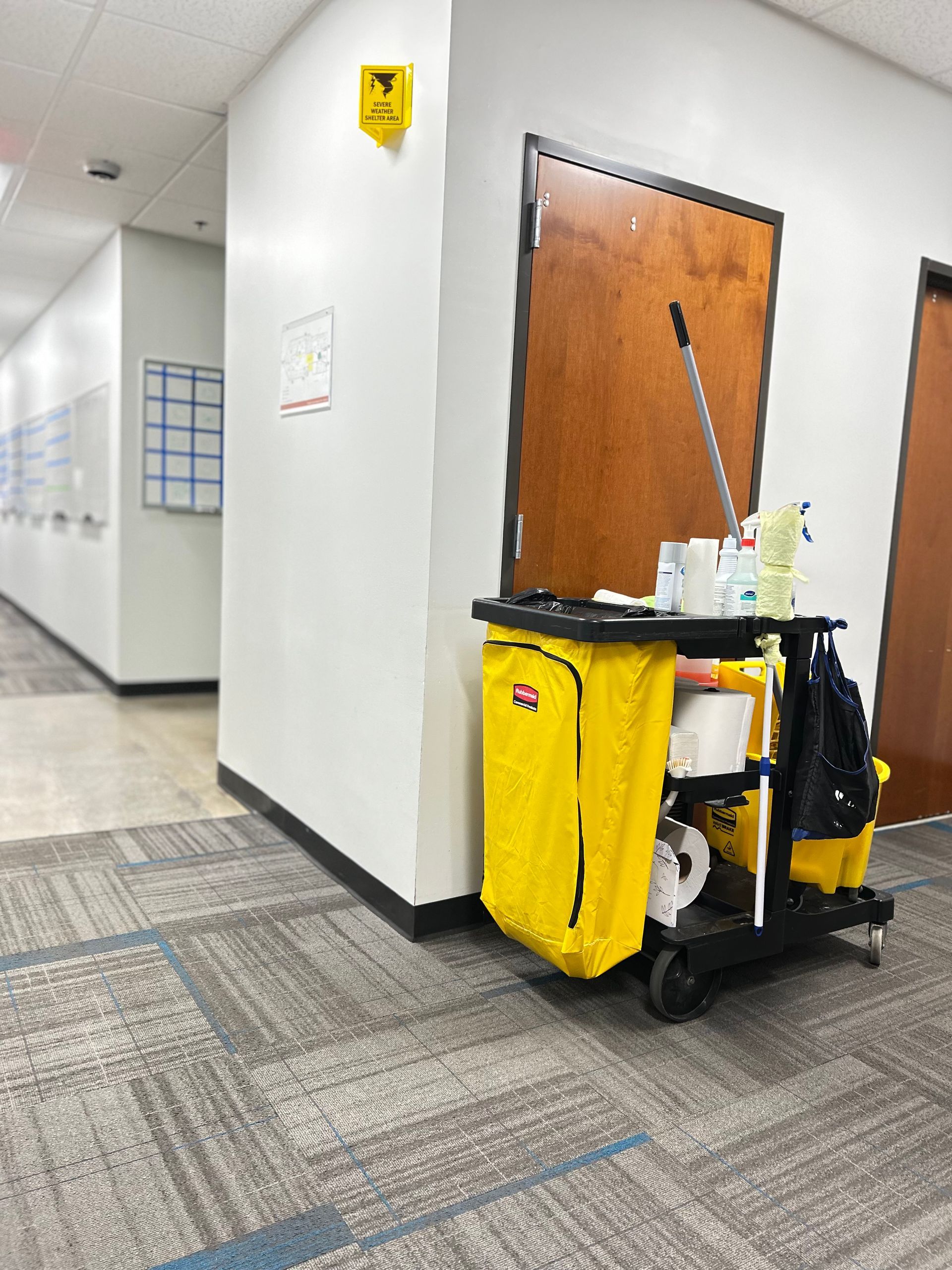 Person in yellow uniform pushing a cleaning cart along a wall in a room.