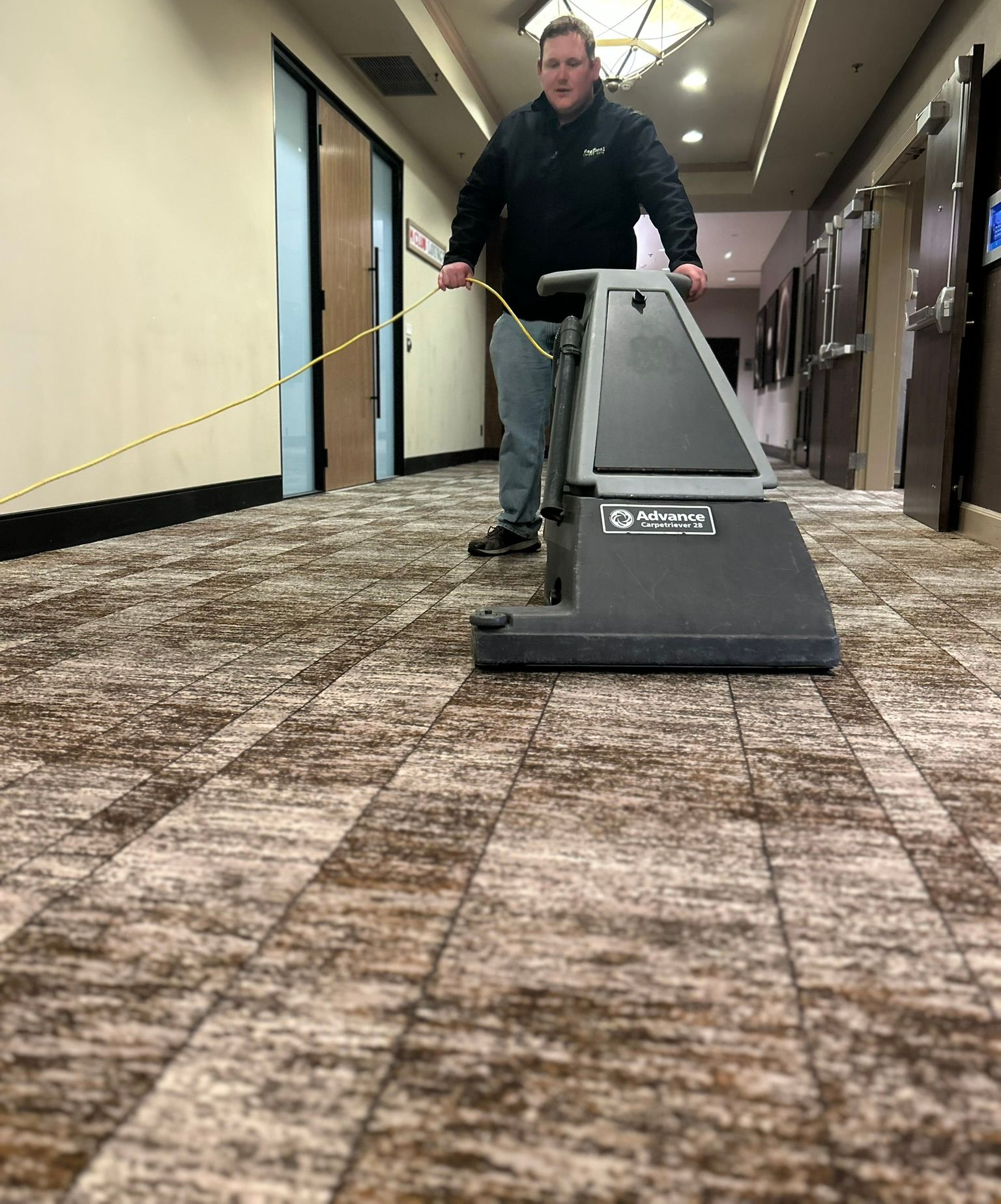 Modern office workspace with desks, chairs, and computers. The room has large windows and a gray patterned carpet.