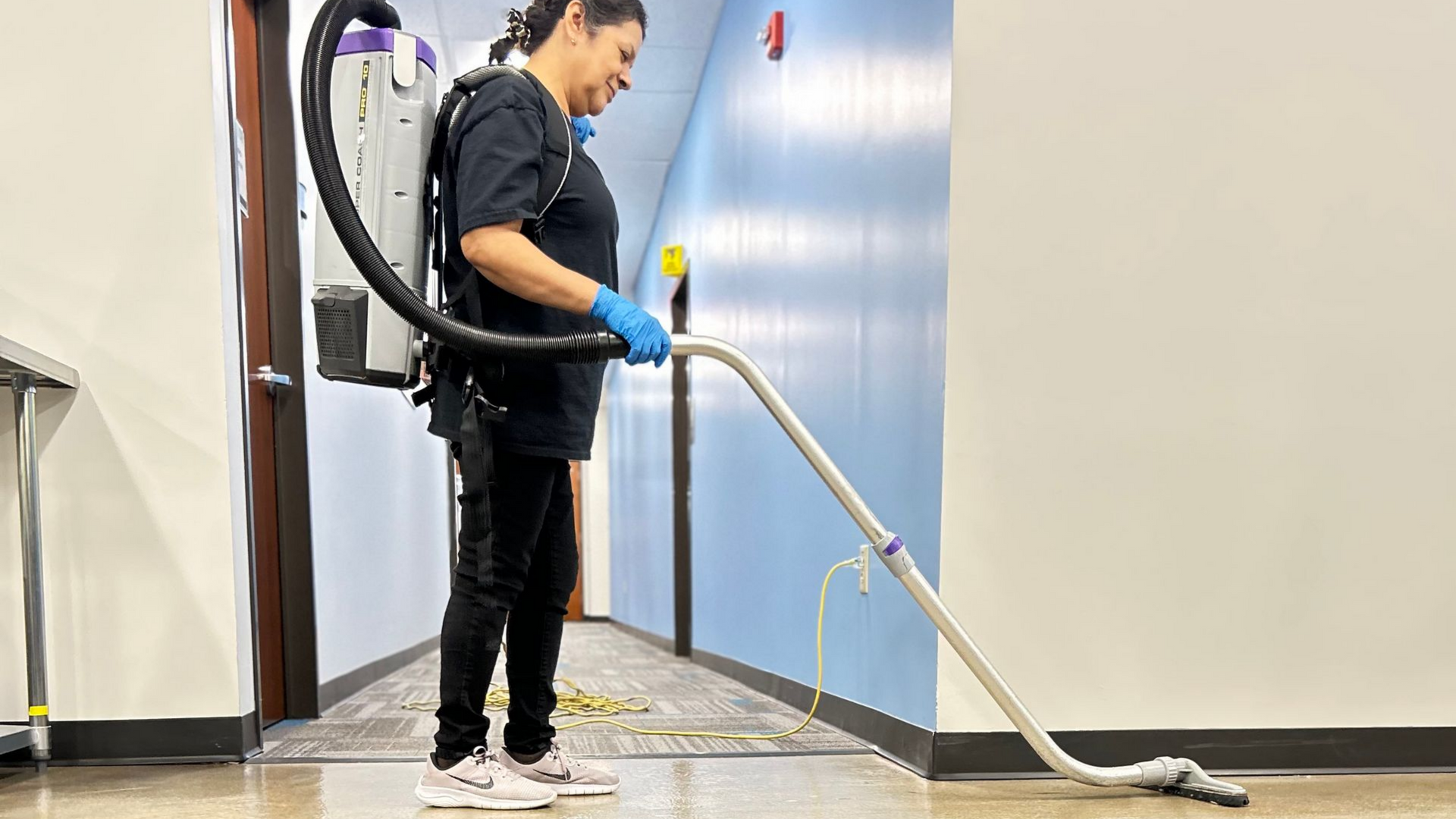 Person using a floor buffer on a dark floor in a room. Another person walks in the background.