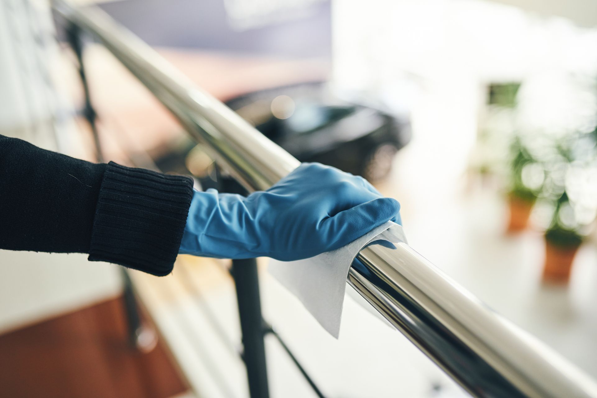 A gloved hand wipes a metal handrail with a cleaning wipe indoors.