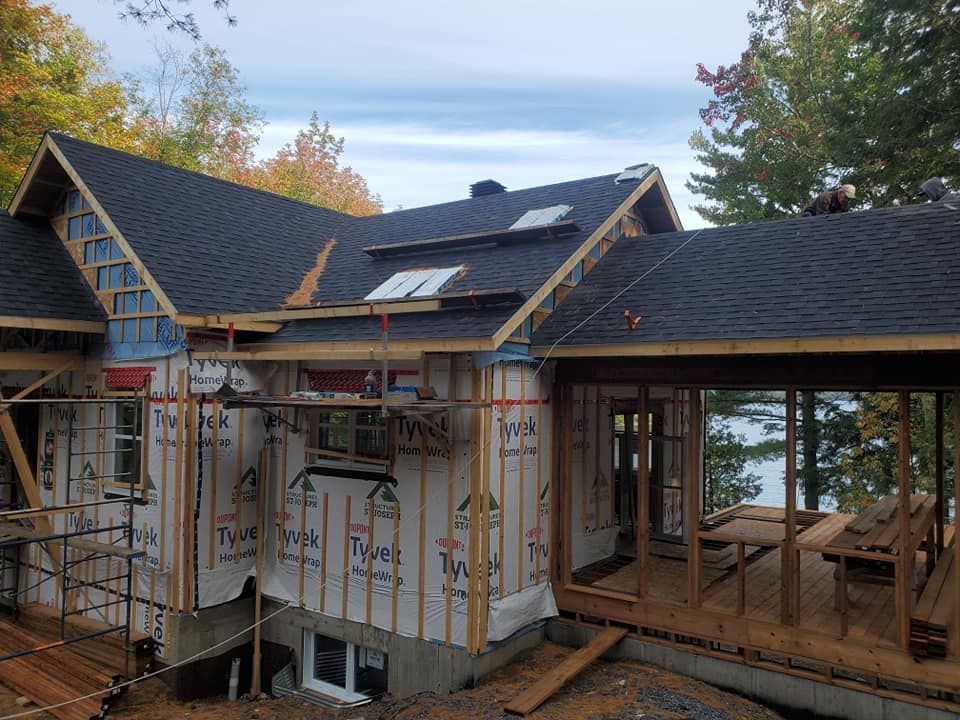 Maison en construction avec toit foncé, ossature bois et revêtement bleu. Terrasse en bois donnant sur l'eau.