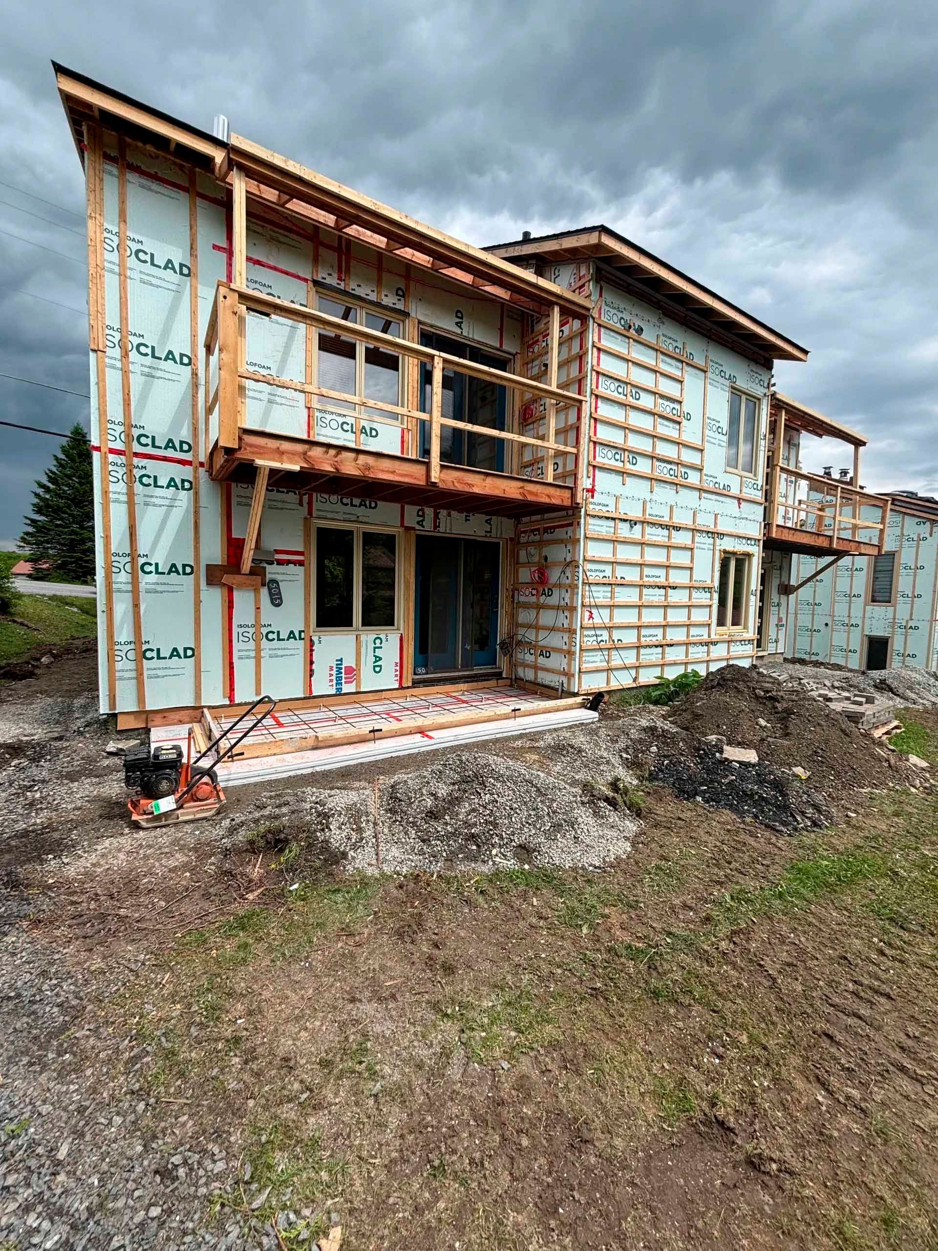 Maison à deux étages en construction, avec ossature bois, balcon et pare-vapeur. Ciel couvert en arrière-plan.
